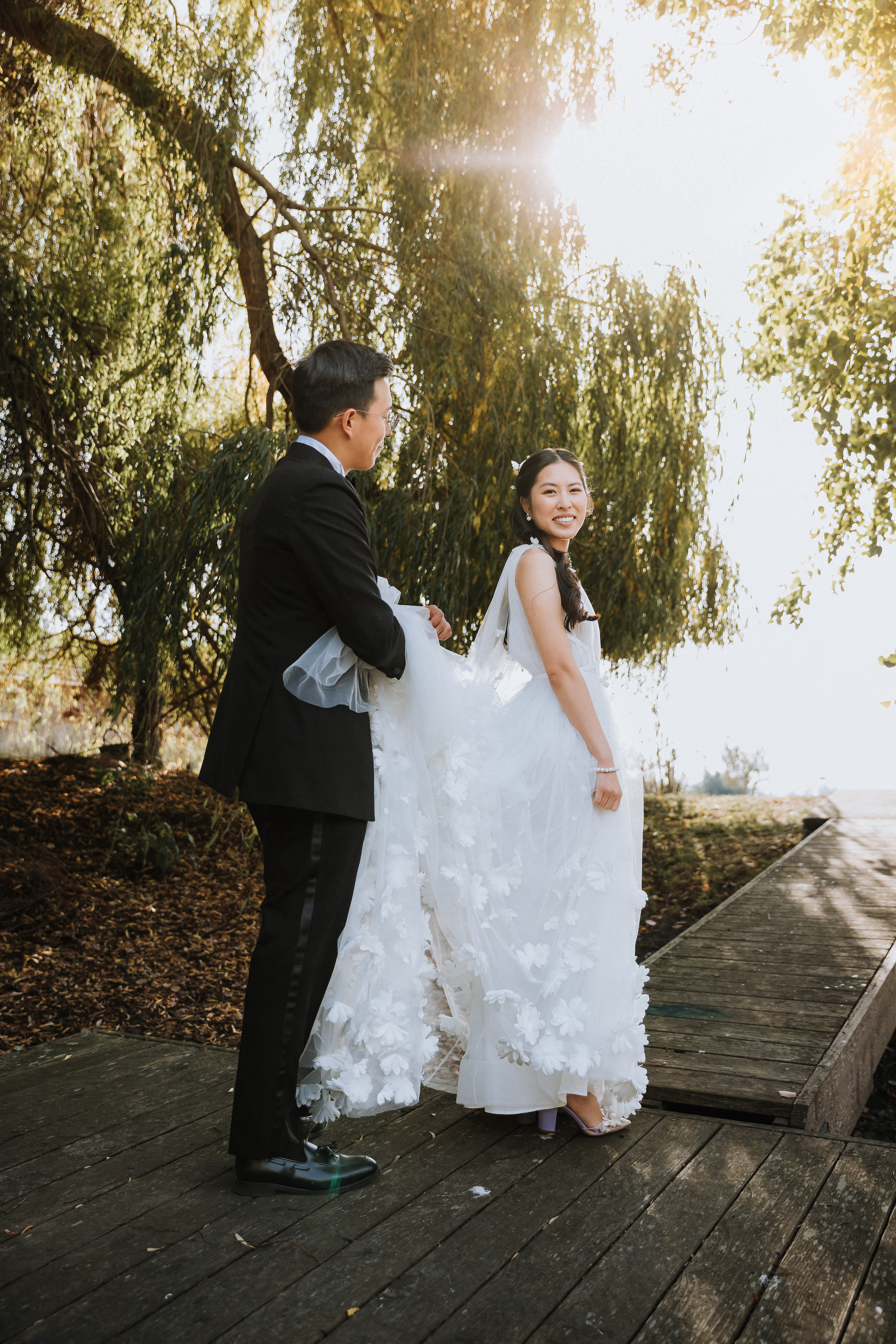 Groom looking at bride while holding her gown's train on a dock near the lake under the trees. Bride is smiling for the photo.