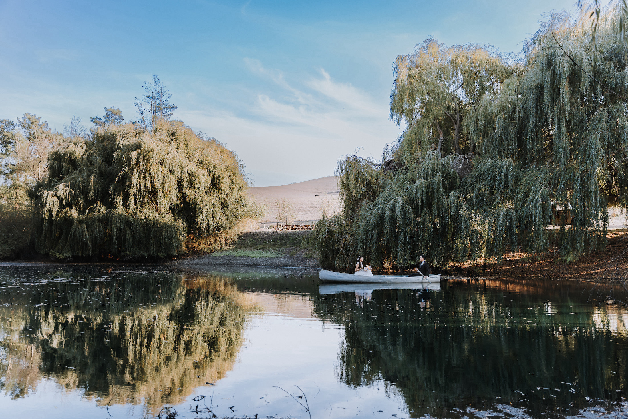 Wide shot of bride and groom on a row boat on the lake under the willow trees.