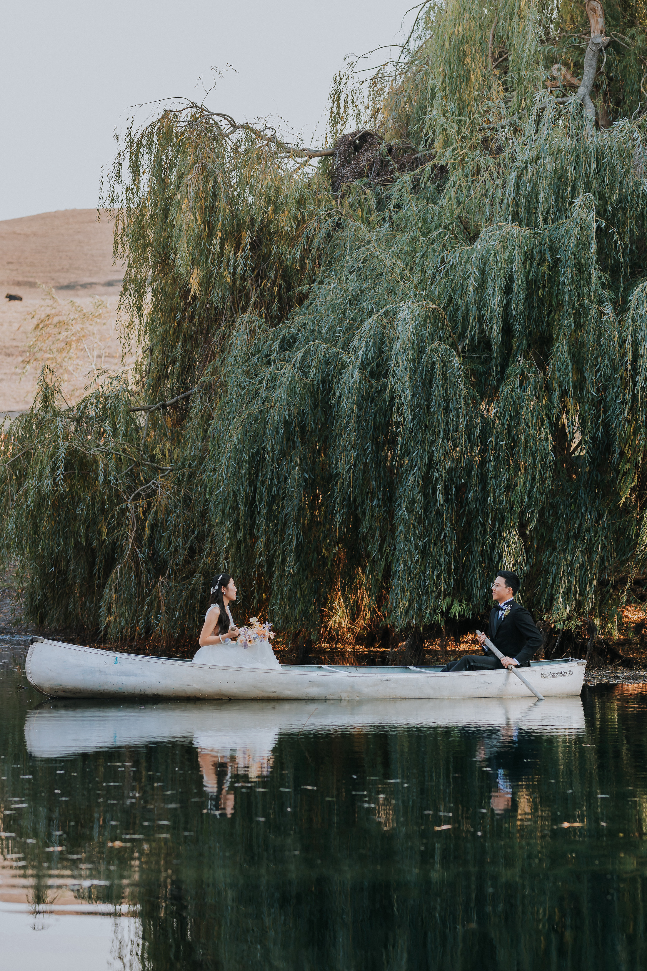 Bride and groom on a row boat on the lake under the willow trees. The groom is rowing on one end and the bride is sitting with her bouquet in hand on the other end.