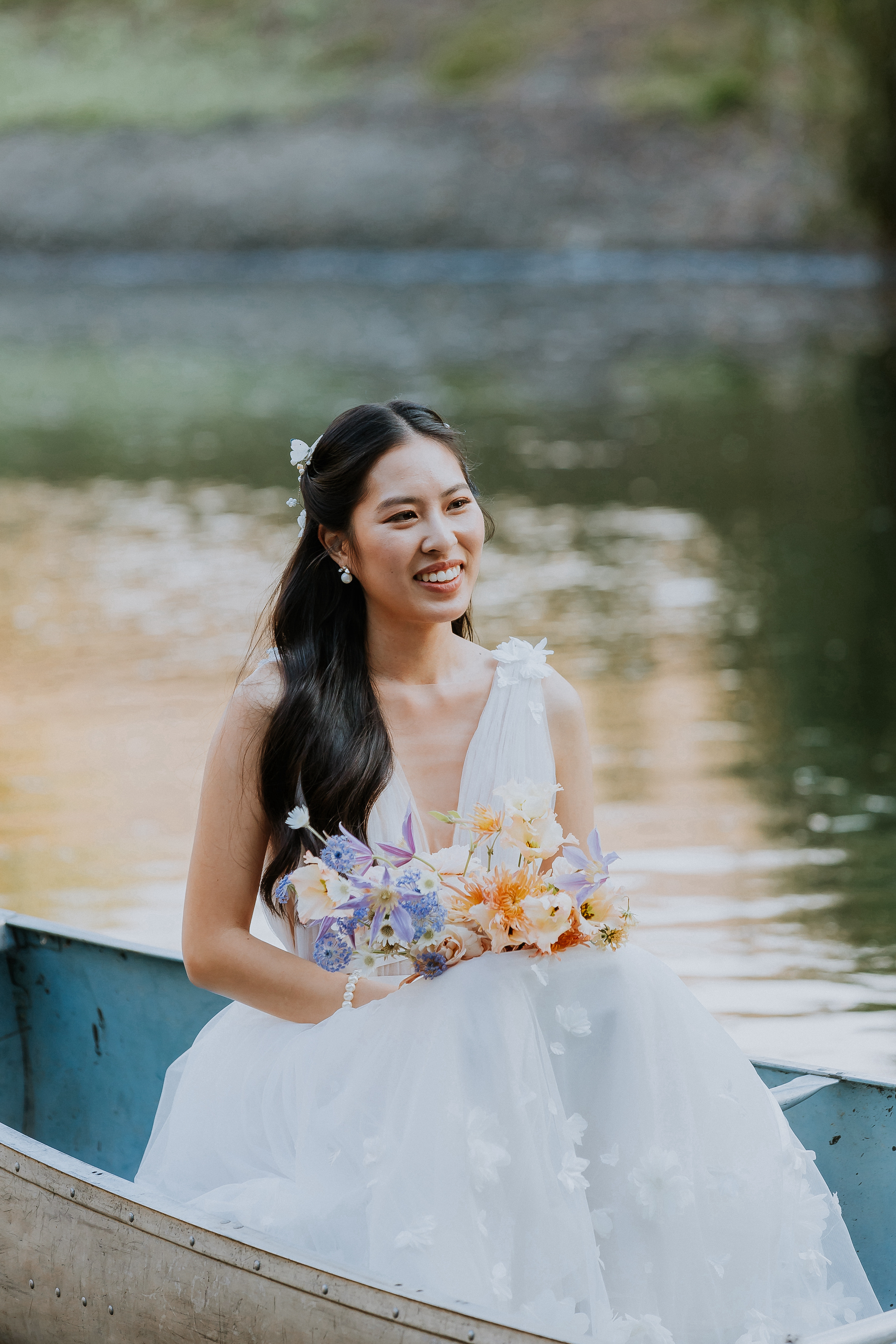 Bride holding bouquet and smiling while sitting in a rowboat on a lake.