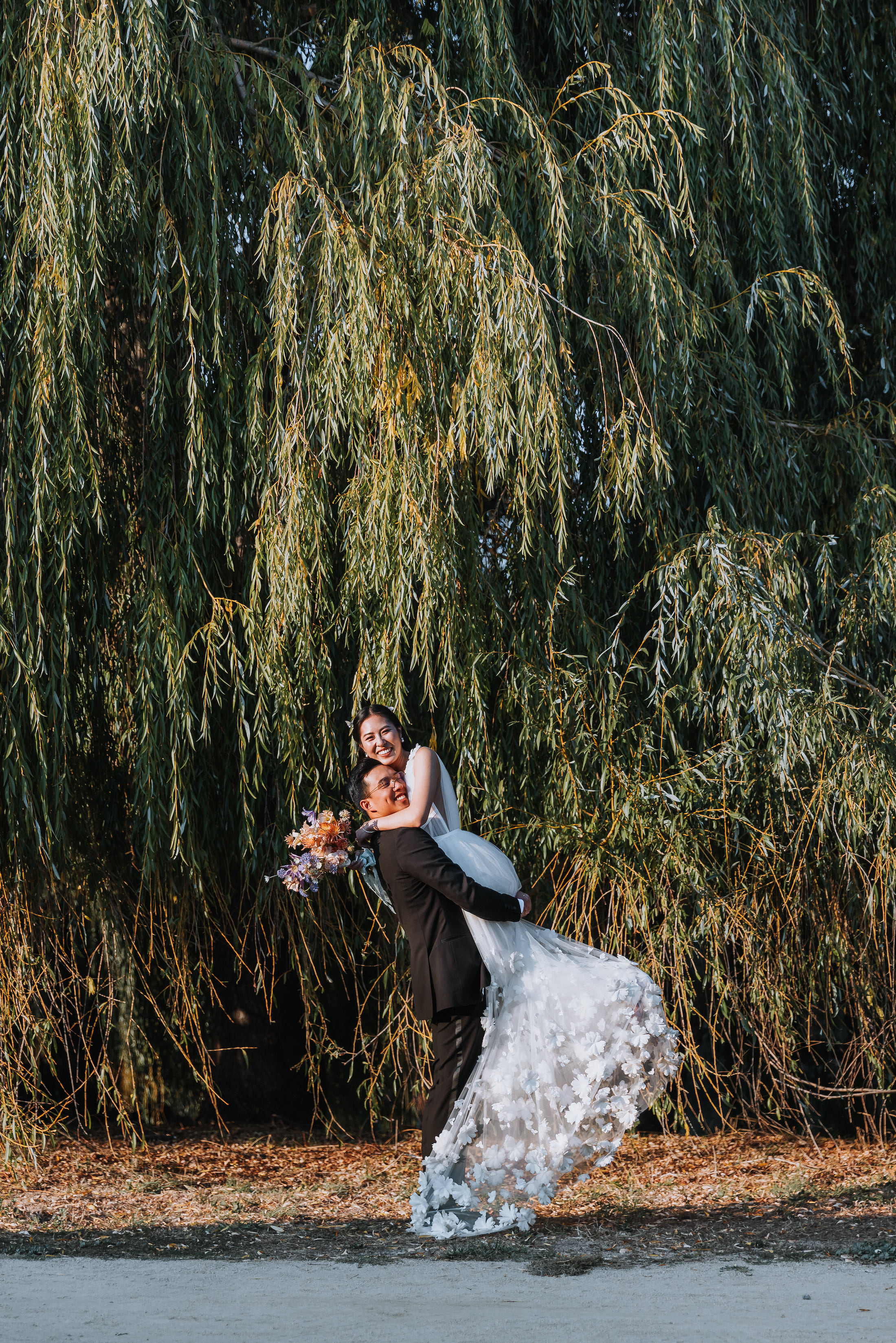 Groom lifting bride up in a large embrace in front of willow trees while they smile.
