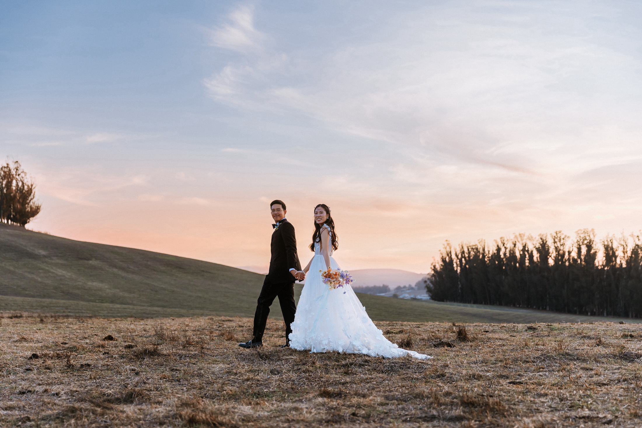Bride and groom smiling and holding hands in a field during sunset.