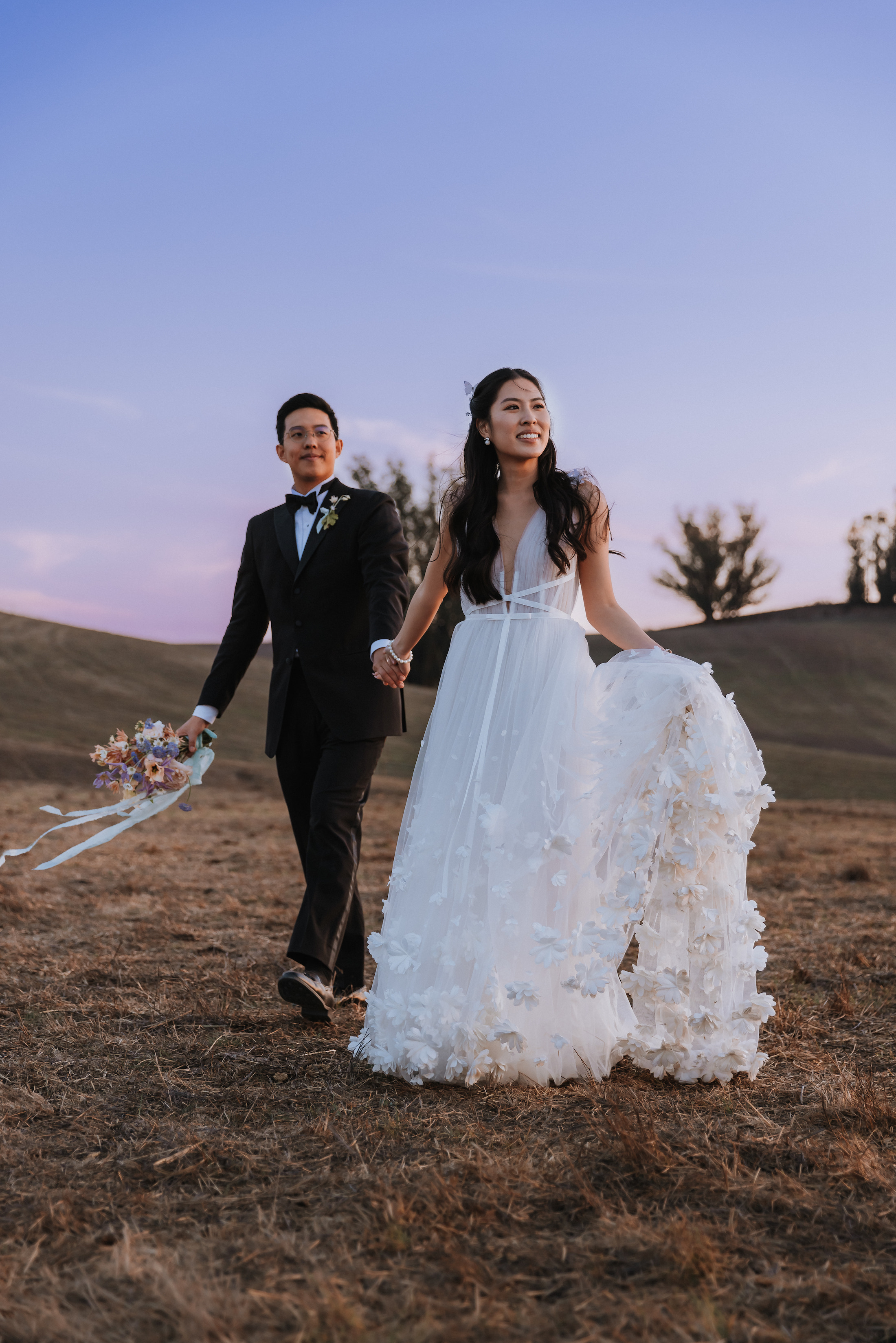 Bride and groom smiling and holding hands while walking in a field during sunset.