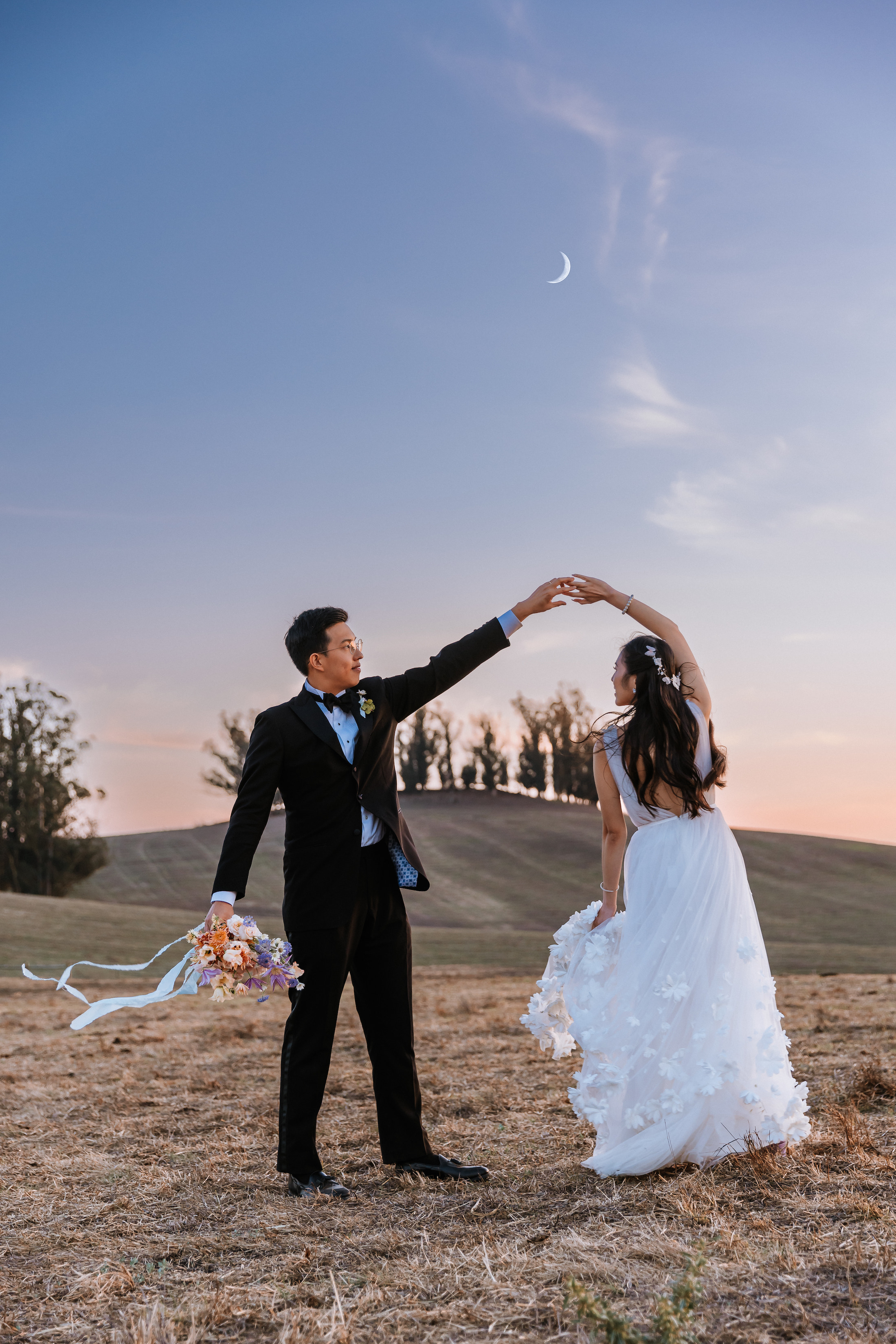 Bride and groom dancing in a field during sunset with the crescent moon above.