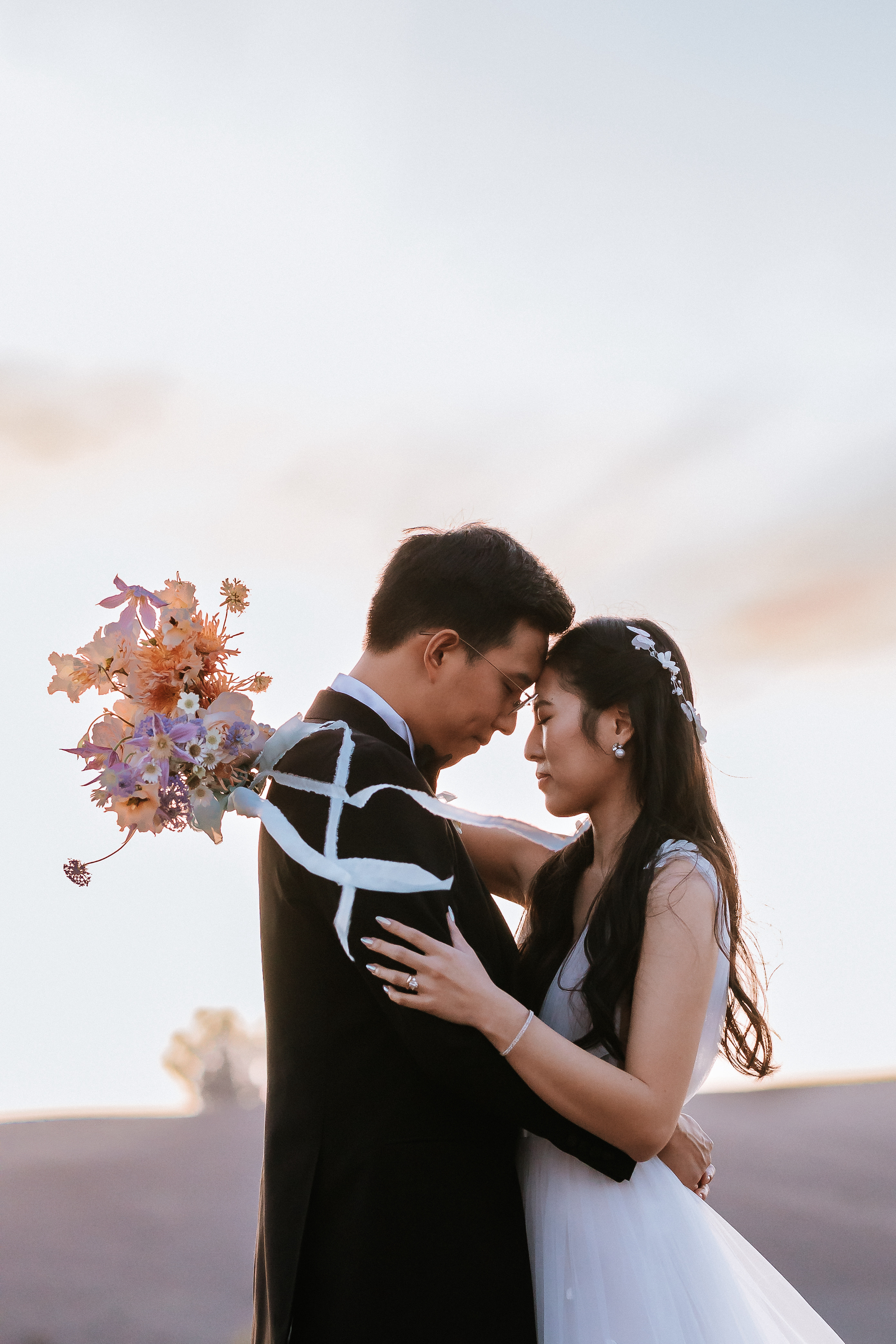 Bride and groom embracing while touching foreheads with eyes closed in a field during sunset.