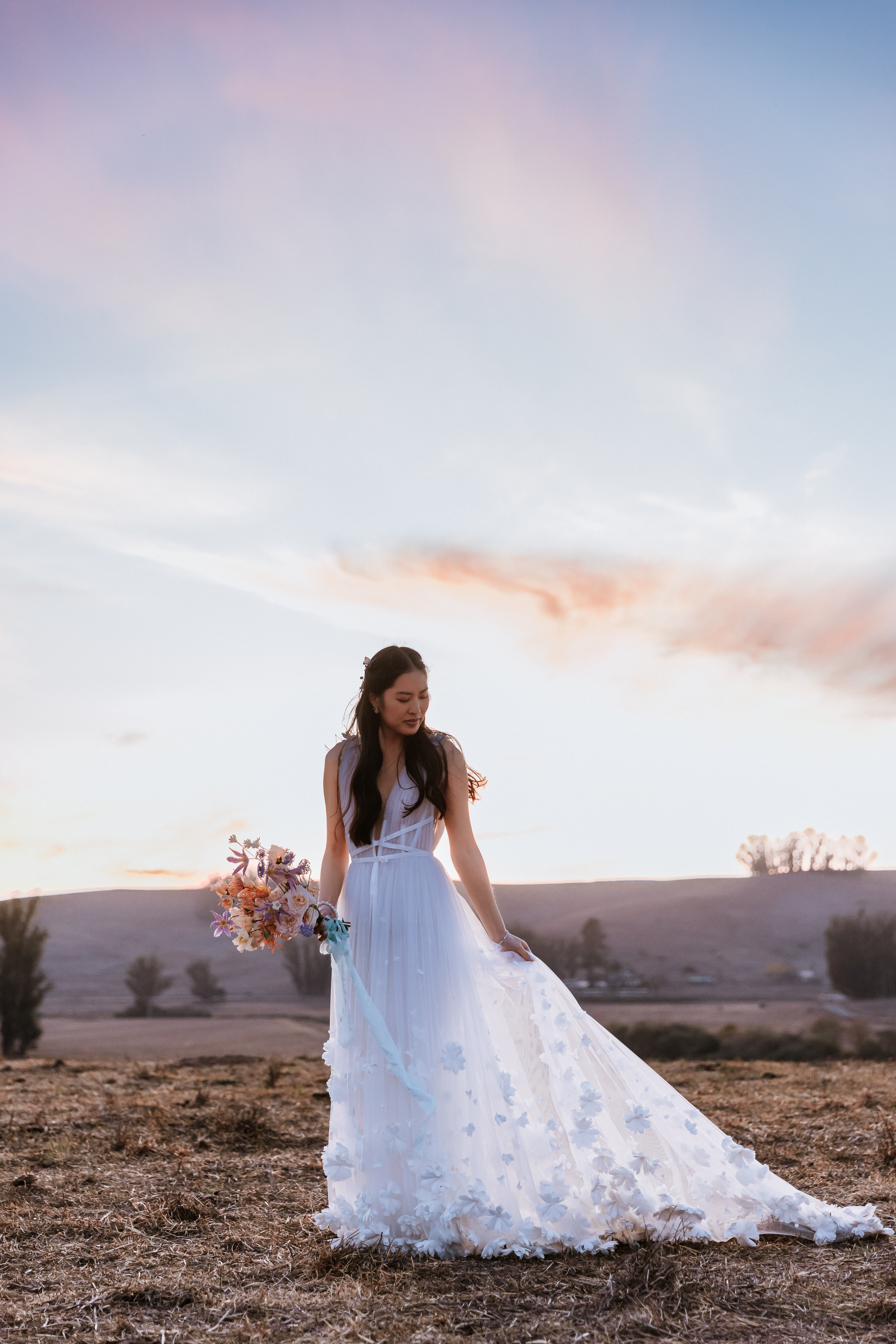 Bride alone holding her bouquet in one hand and her skirt in the other hand while in a field during sunset.