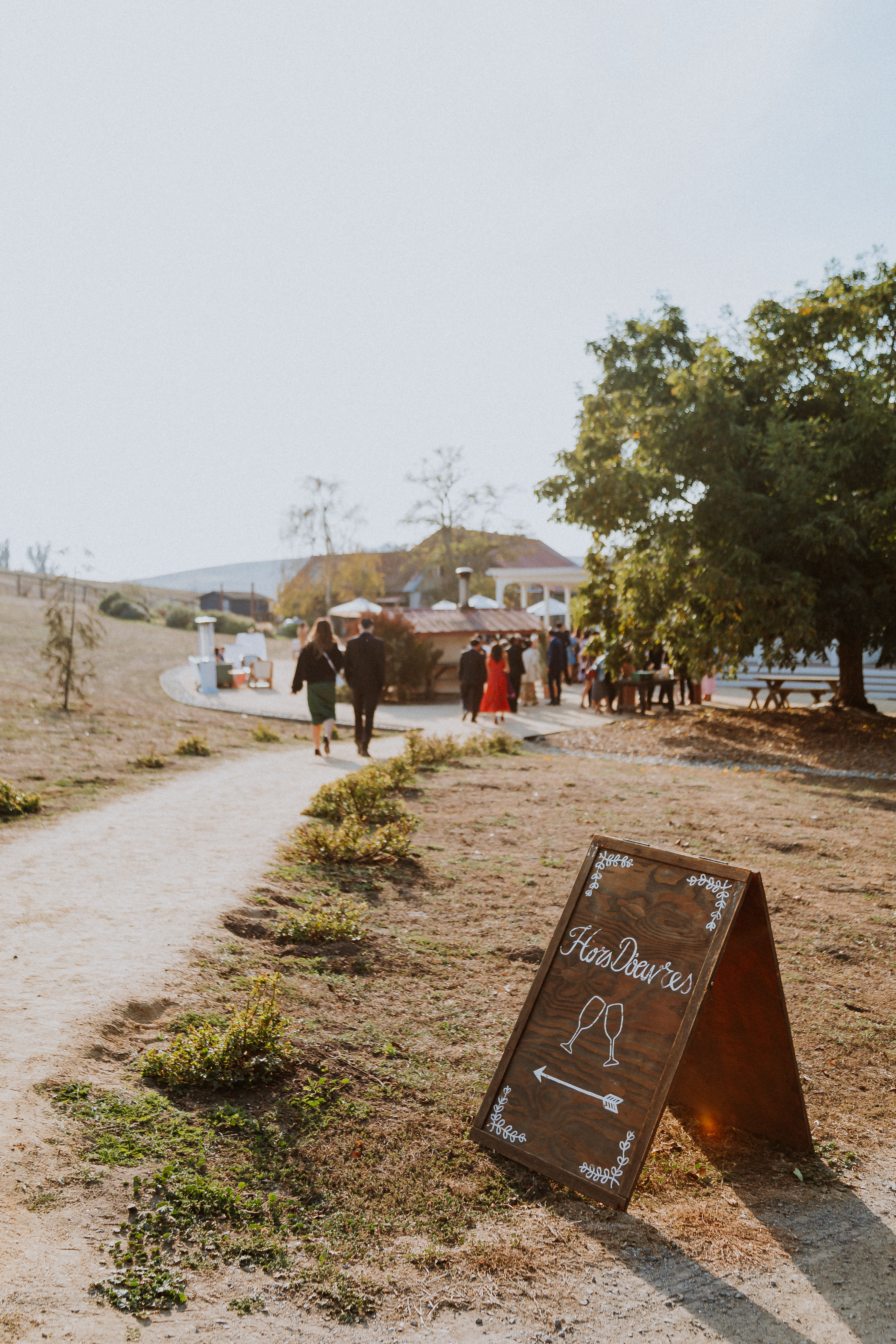 Wedding sign on the ground that reads "hor dourves" and has and illustration of two glasses clinking and an arrow that points to cocktail hour as people walk past in the background.