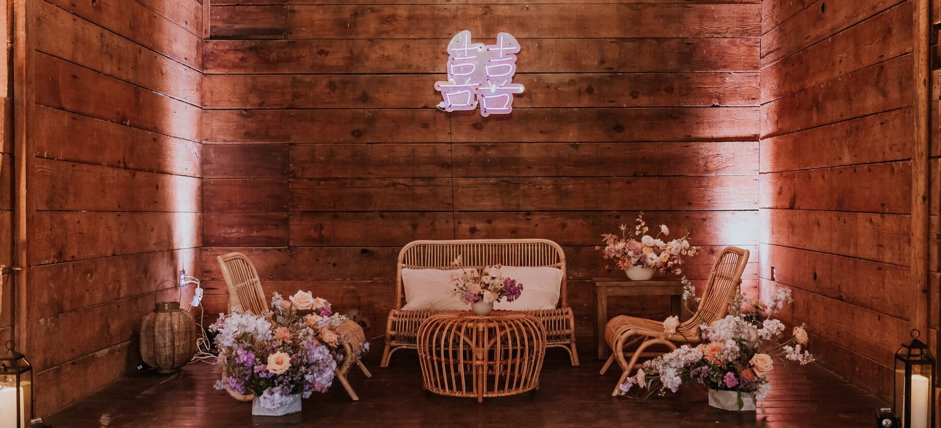 Elegant wedding lounge designed by Two Perfect Events, featuring rattan furniture, soft floral arrangements, and a glowing double happiness neon sign against a rustic wooden backdrop. A beautifully curated space for unforgettable wedding experiences and luxury event design in the San Francisco Bay Area and beyond.