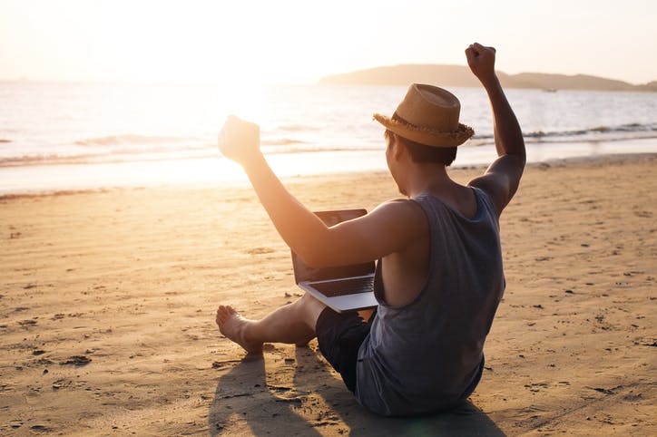 Young man sitting on beach at sunset in shorts, vest and hat with laptop on knees and arms aloft