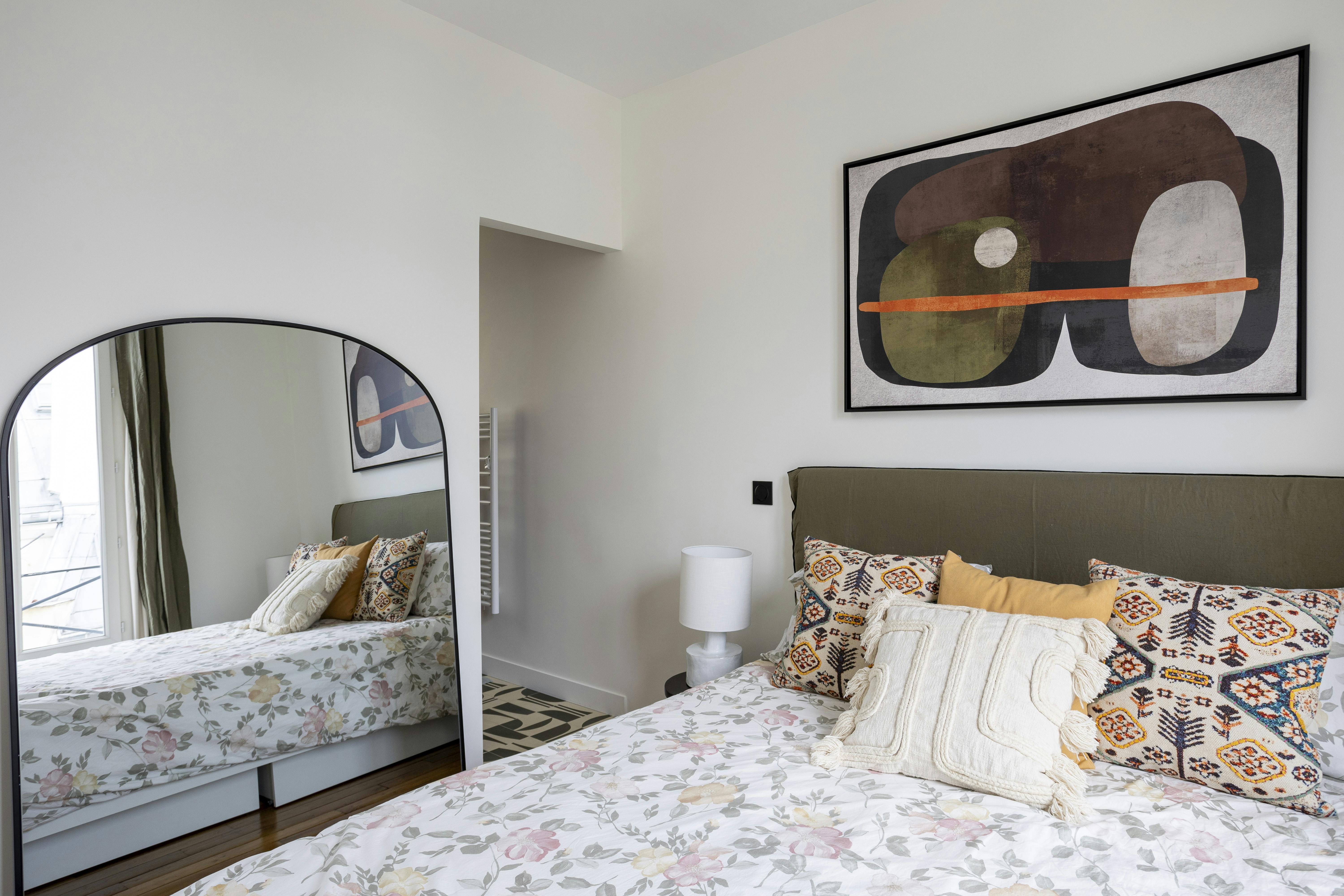 Chambre cosy avec tête de lit kaki, coussins graphiques et grand miroir arrondi.