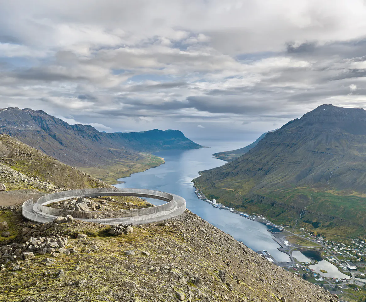 A circular viewing platform overlooks Seyðisfjörður from a mountainside high above the fjord. Steep mountains rise on either side of the water, with patches of snow, drifting mist, and rugged volcanic terrain creating a dramatic East Iceland 