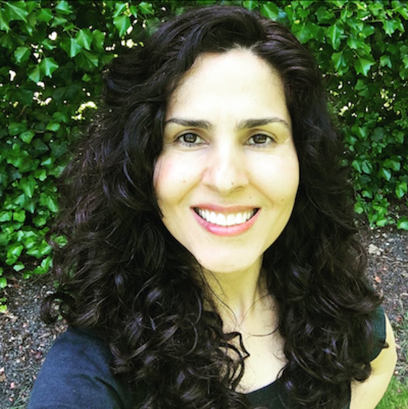 Photo of the author, with curly black hair, flowing past the shoulders with a green leaf background.