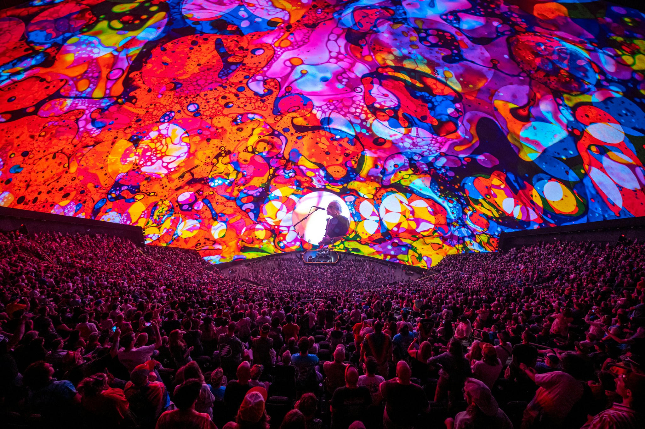 dead and company at sphere, the crowd watch Bob Weir sing and play guitar, he is surrounded by colourful psychedelic patterns