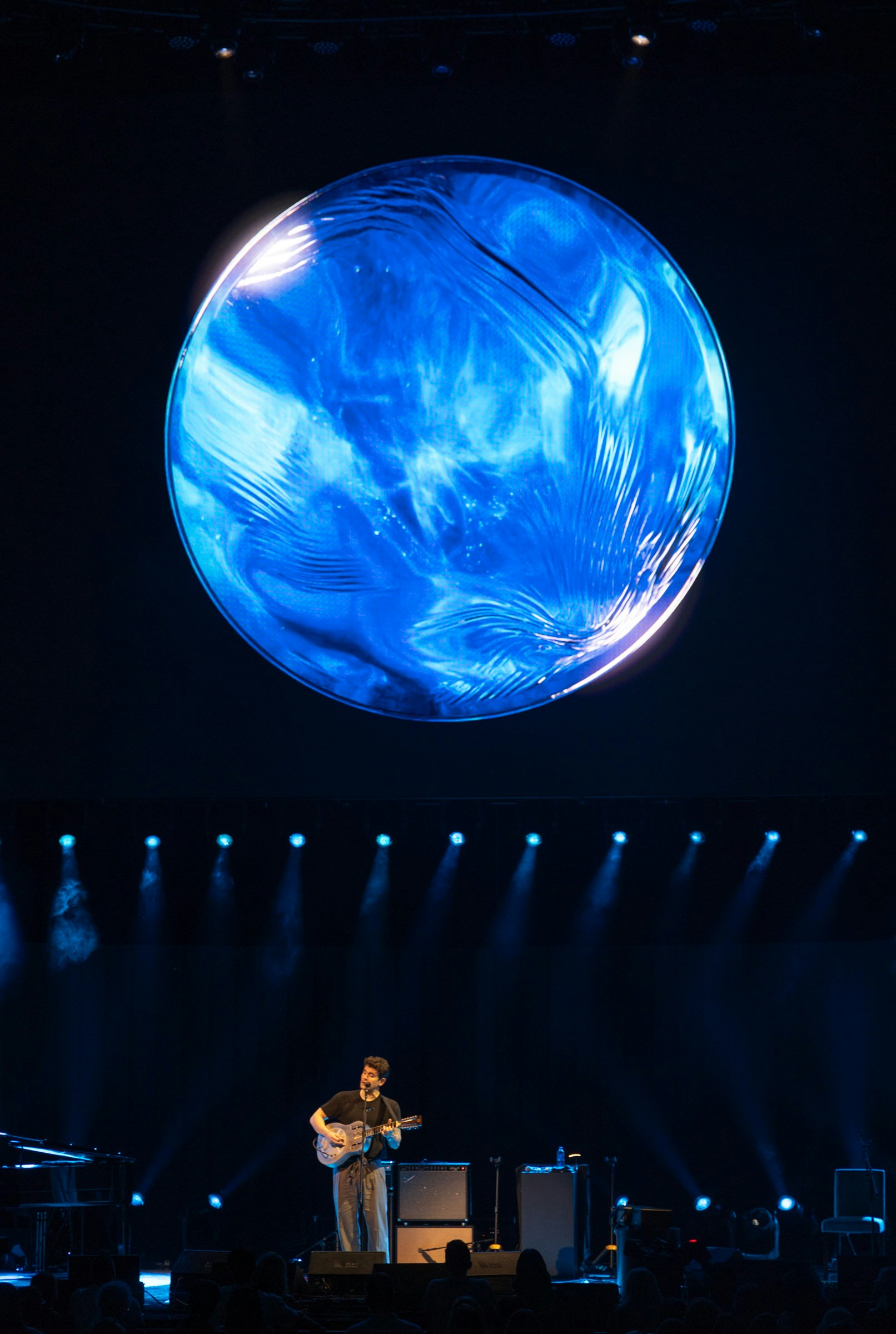 John Mayer stands holding a guitar beneath a blue orb