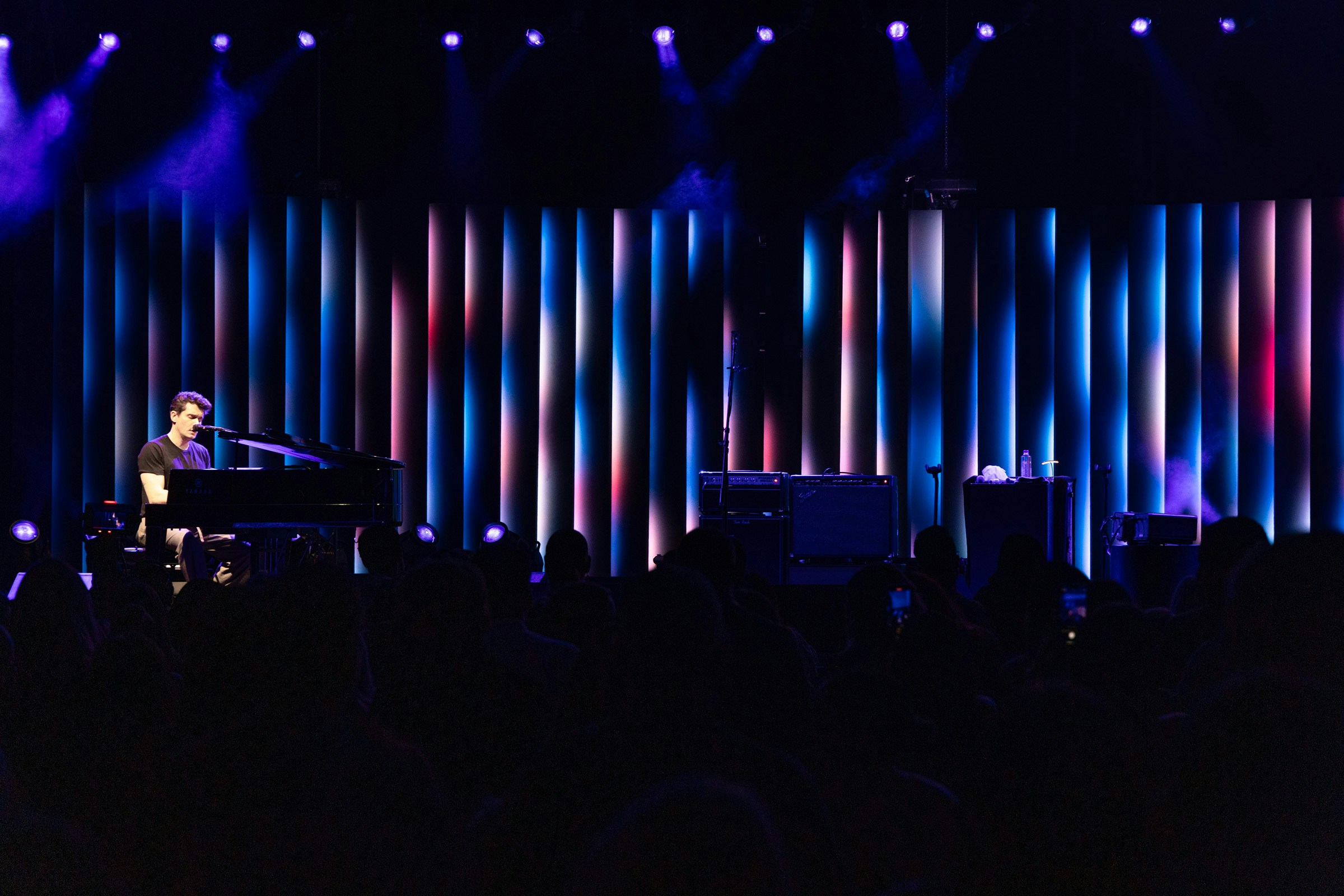 john mayer sits behind a piano on a stage, behind him are vertical slats lit by different coloured lights