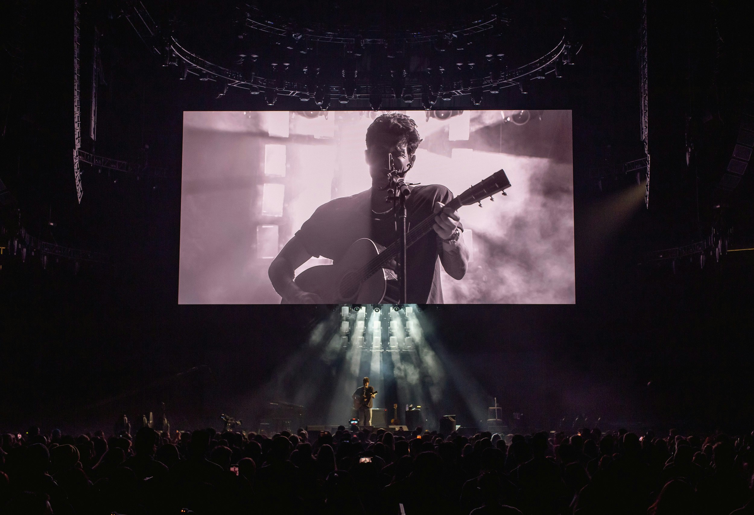 John Mayer stands holding a guitar on a stage back lit by shafts of light