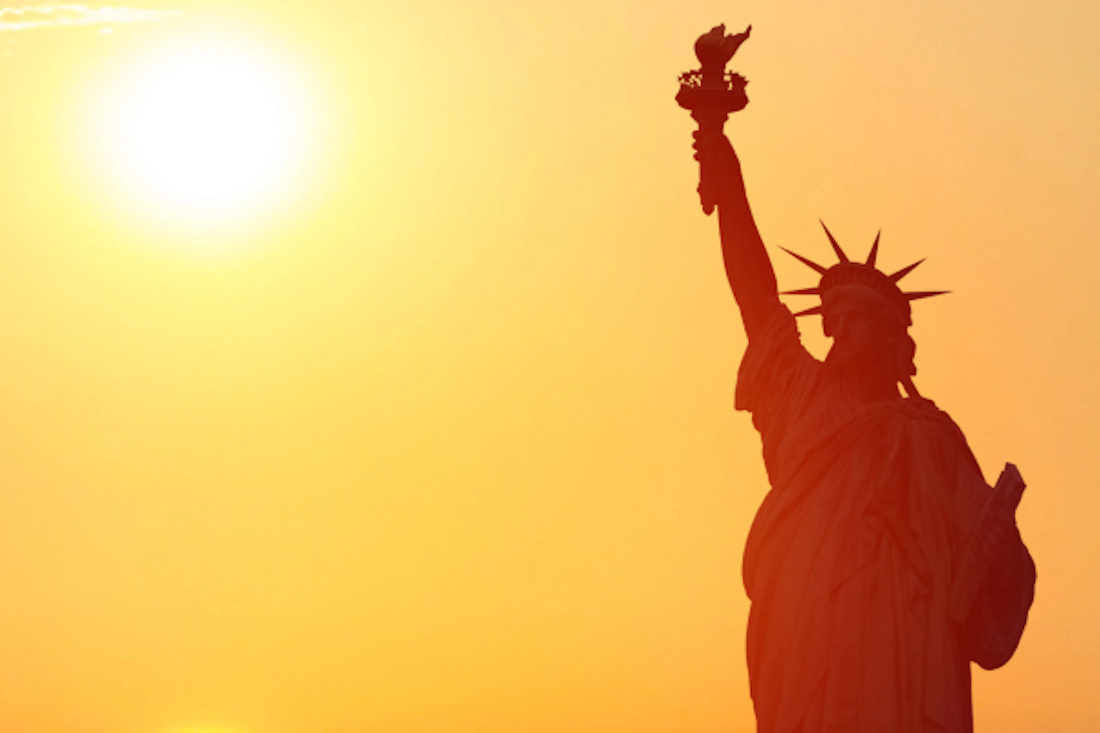the sun sets behind the statue of liberty, casting it into a silhouette 