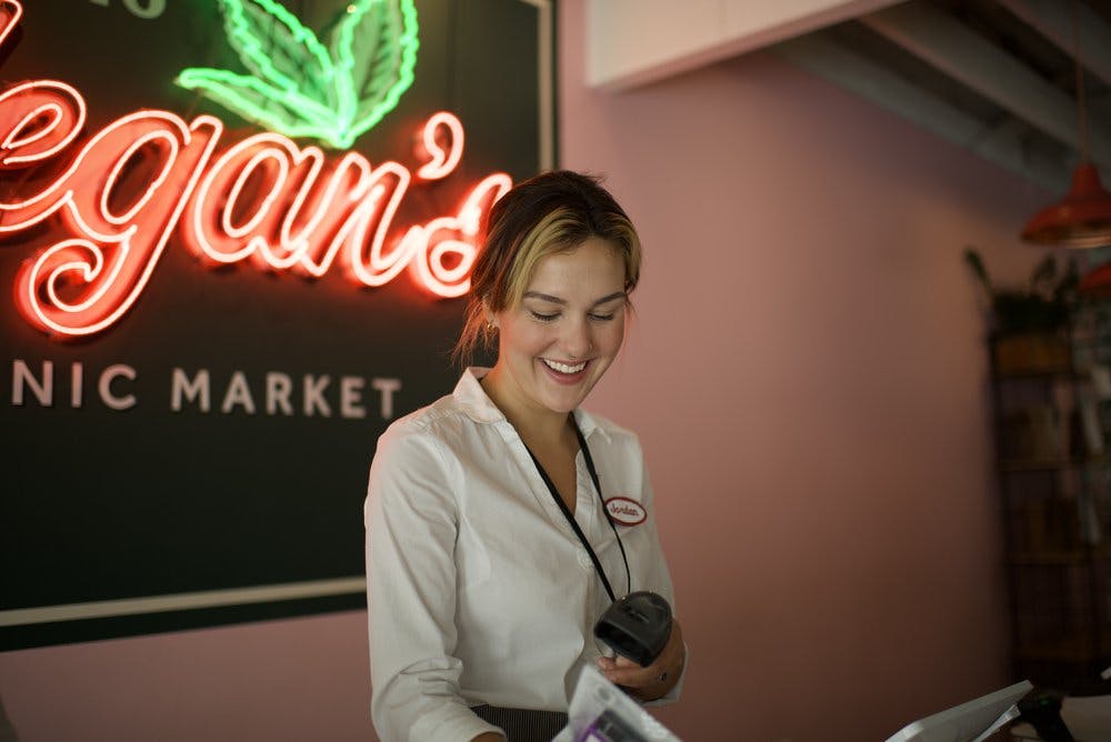A smiling budtender scans product in front of a neon sign that says Megan's Organic Market