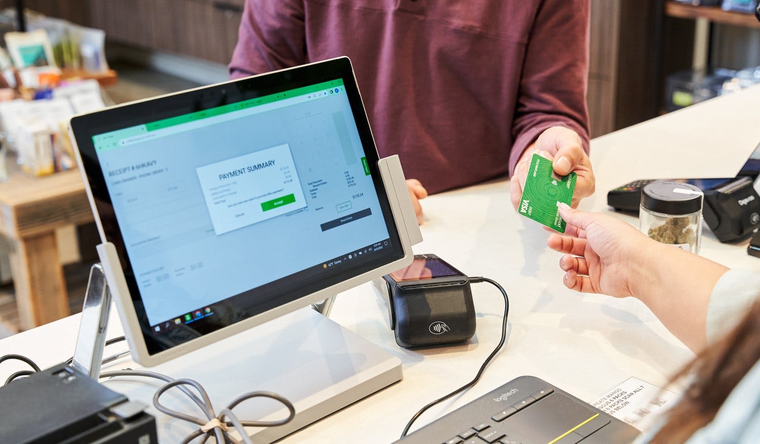 Two hands reach over a counter with glass jars of cannabis, a point of sale system shows Payment Summary