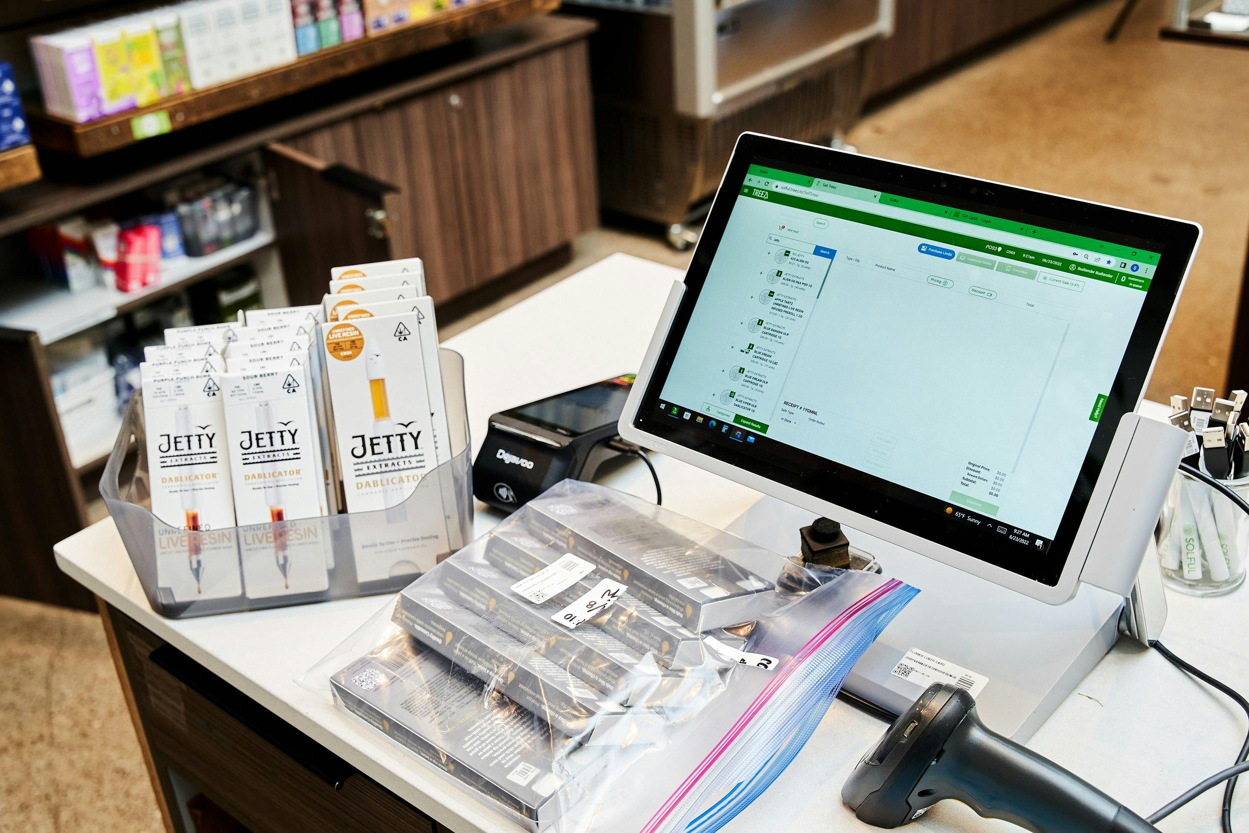 A point of sale sits on a counter, with cannabis products like vape cartridges and vape batteries around. There is a scanner and a cashless payments terminal.