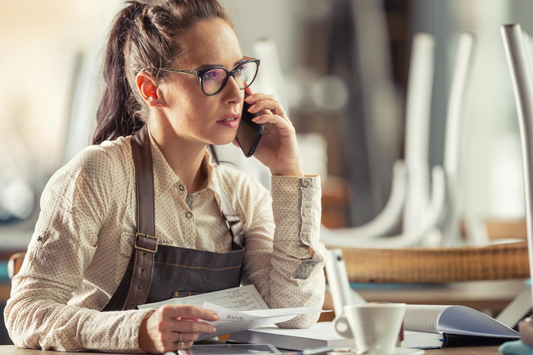 A business owner sits at their desk, with a phone to their ear and a bill in their other hand. they wear a face of concern.