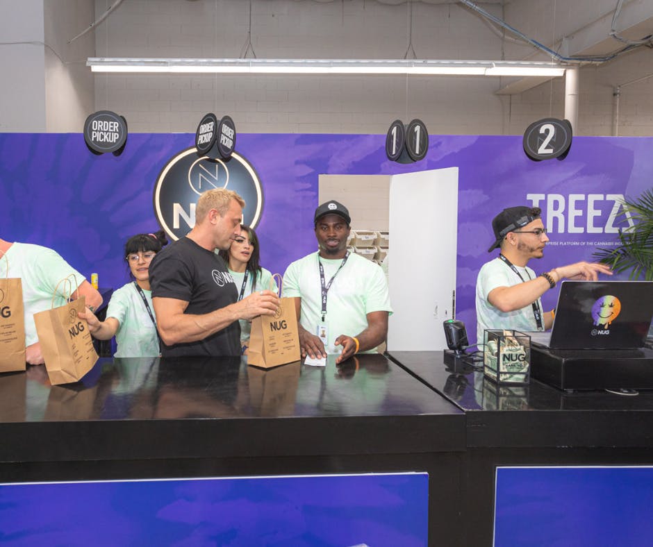 Behind the counter of a dispensary are several budtenders serving clients at the Hall of Flowers Dispensary powered by Treez