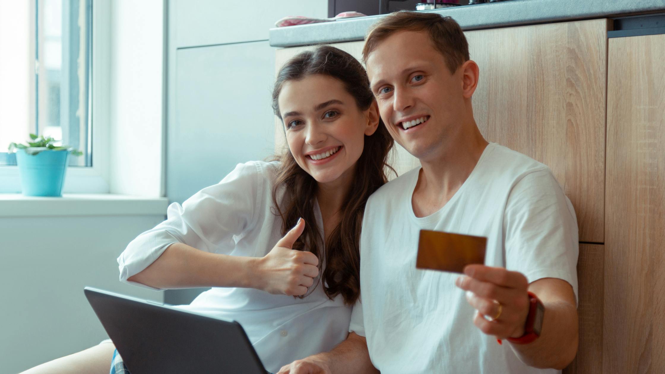 Two smiling people sit on the ground. One holds a credit card up to the viewer, the other has a laptop on their lap and holds a thumbs up to the camera