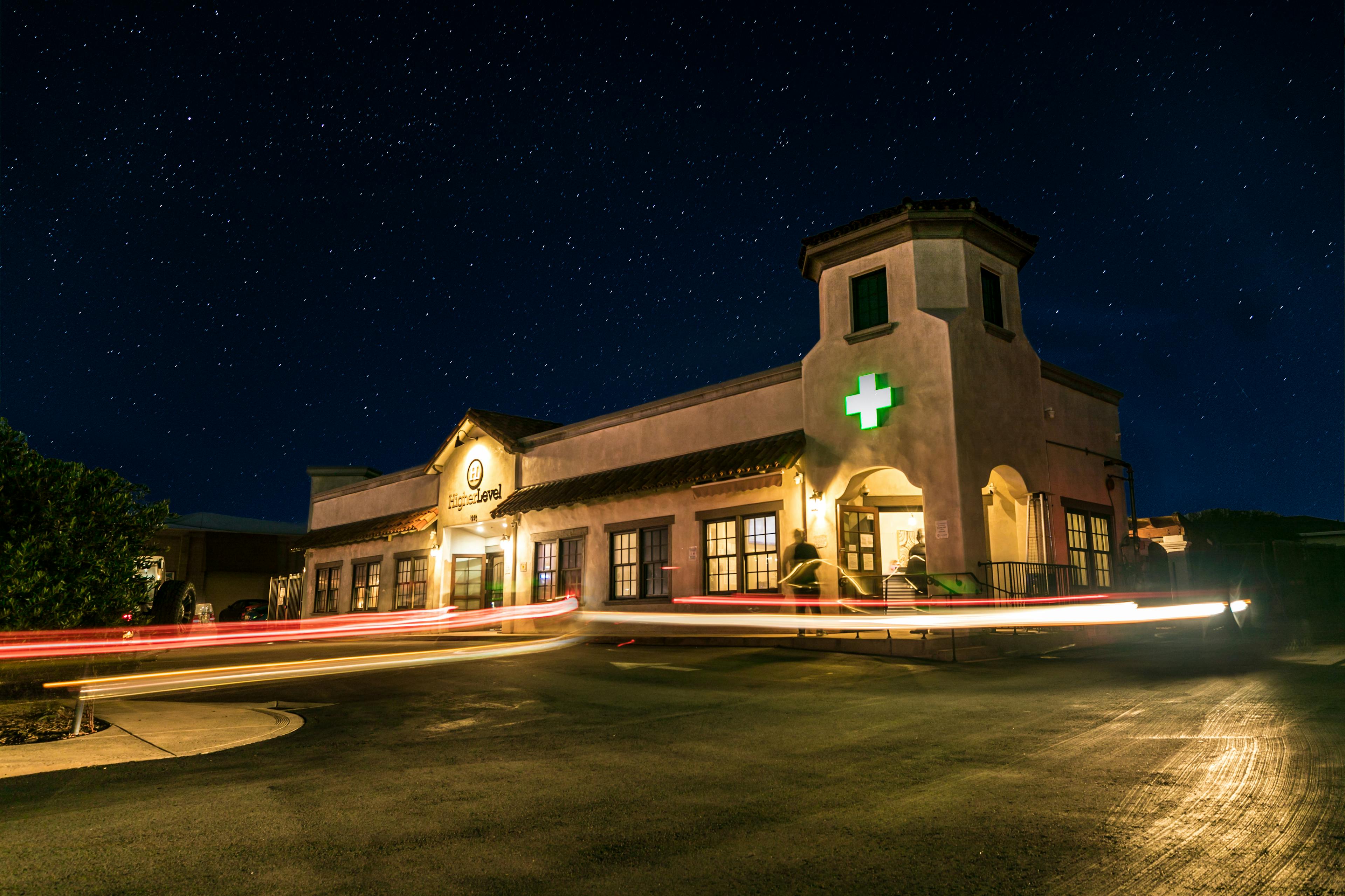 An exterior shot of Higher Level dispensary at night, showcasing the starry sky above and light trails from passing cars, with a green medical cross sign indicating the nature of the business.