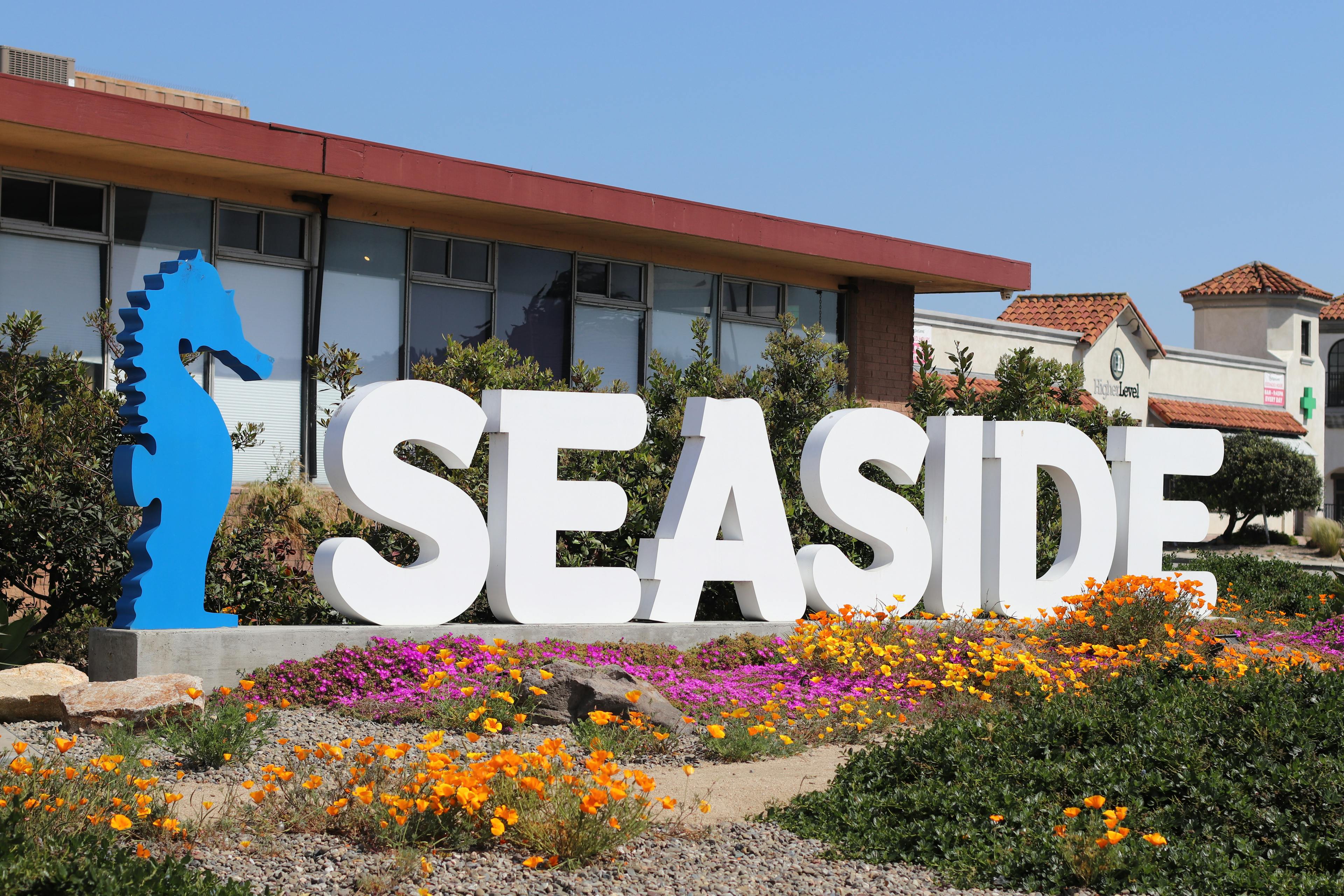 A daytime outdoor image showing a large "SEASIDE" sign with a seahorse silhouette, accompanied by colorful flower beds, suggesting the dispensary's location in a coastal area
