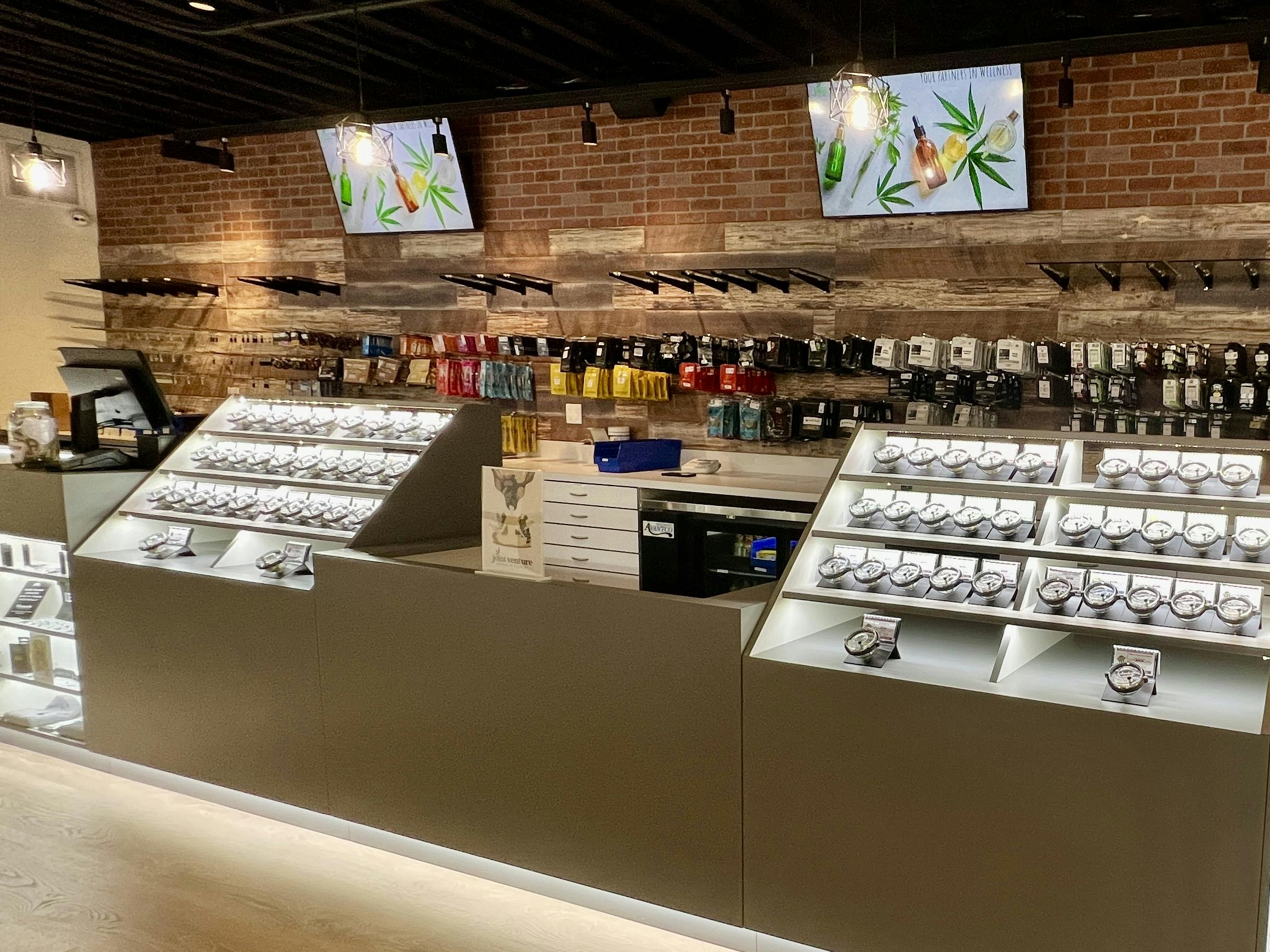 An interior view of a dispensary with empty queues, bright lighting, and a clean, modern aesthetic, displaying products in glass cases and on wall shelves.