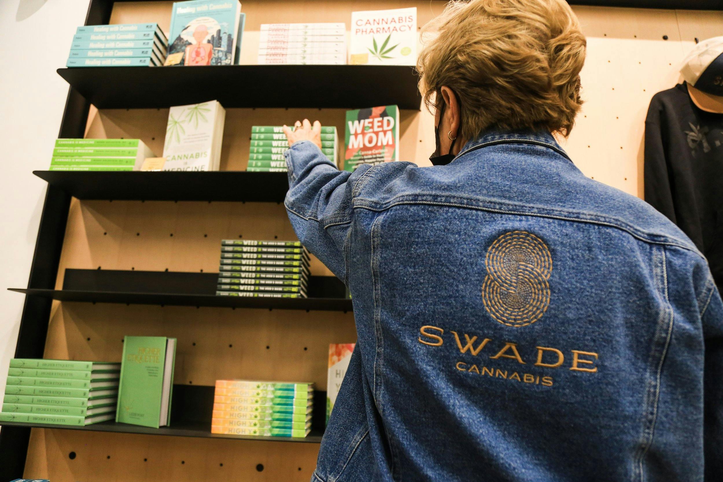A person reaches for a book on display shelves inside a modern dispensary - they are wearing a Swade Cannabis jean jacket and reaching for the book title Weed Mom