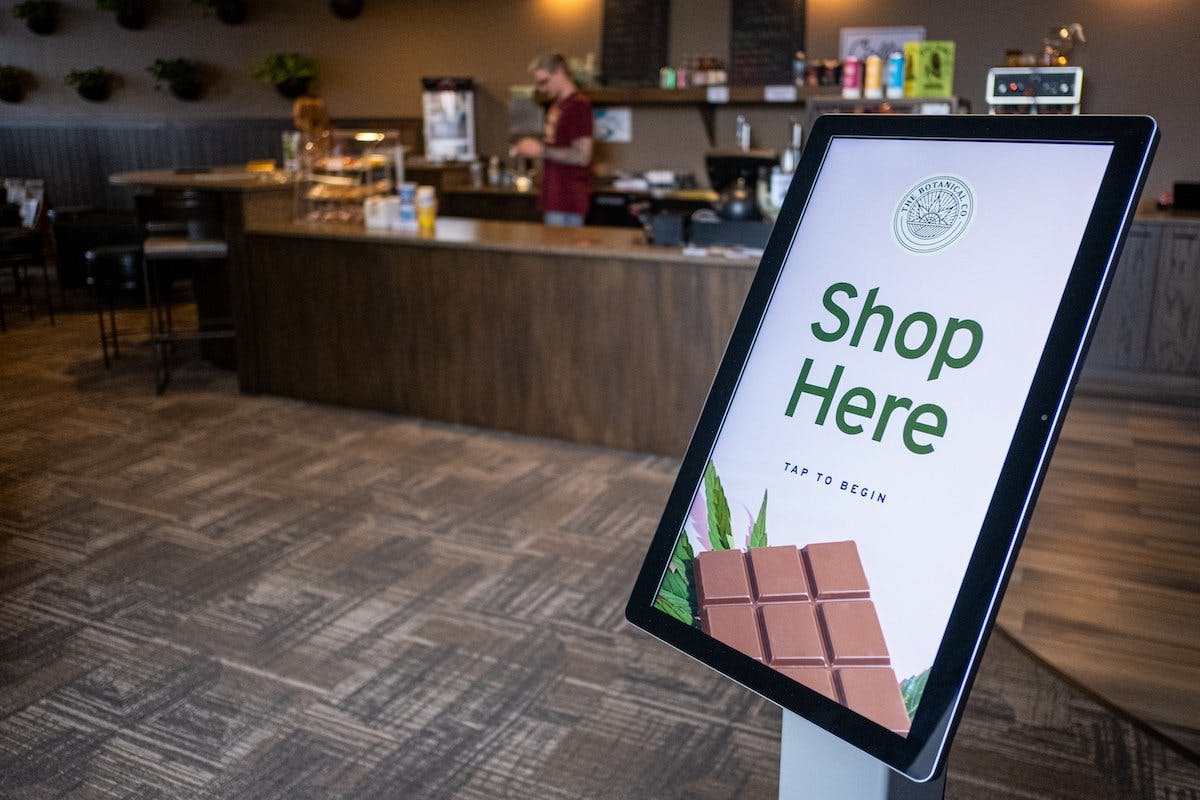 A tablet stands on a pole in front of a check out counter - a chocolate bar and cannabis leaf are displayed below a large message Shop Here and the dispensary's logo