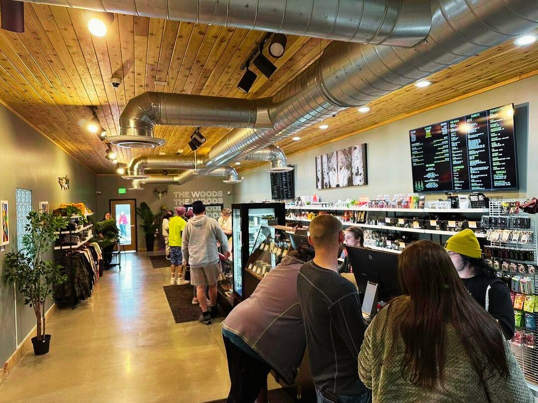 A dispensary sales floor is shown - customers check out at the point of sale system, digital signage shows prices and deals, and the shelves are stocked with cannabis products and accessories
