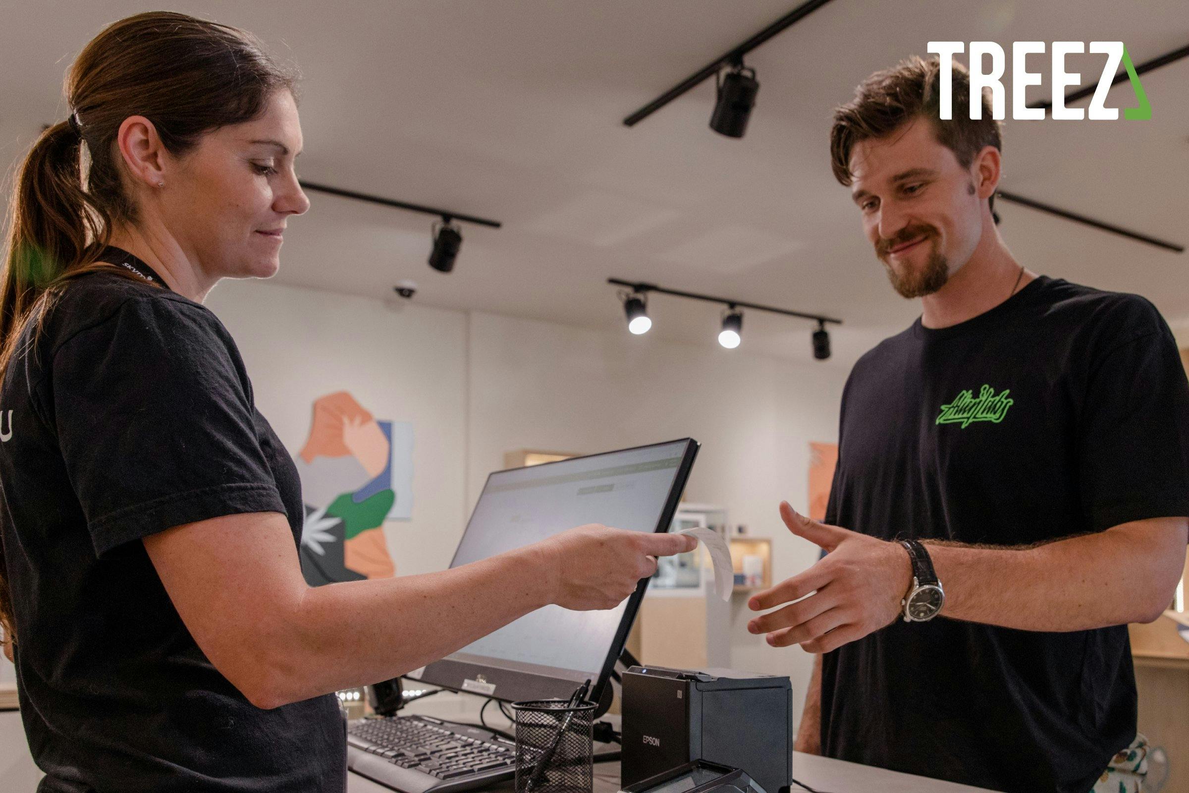 A budtender hands a receipt to a customer after a transaction at a cannabis dispensary. On the counter a computer, keyboard and printer make up a Treez point of sale system