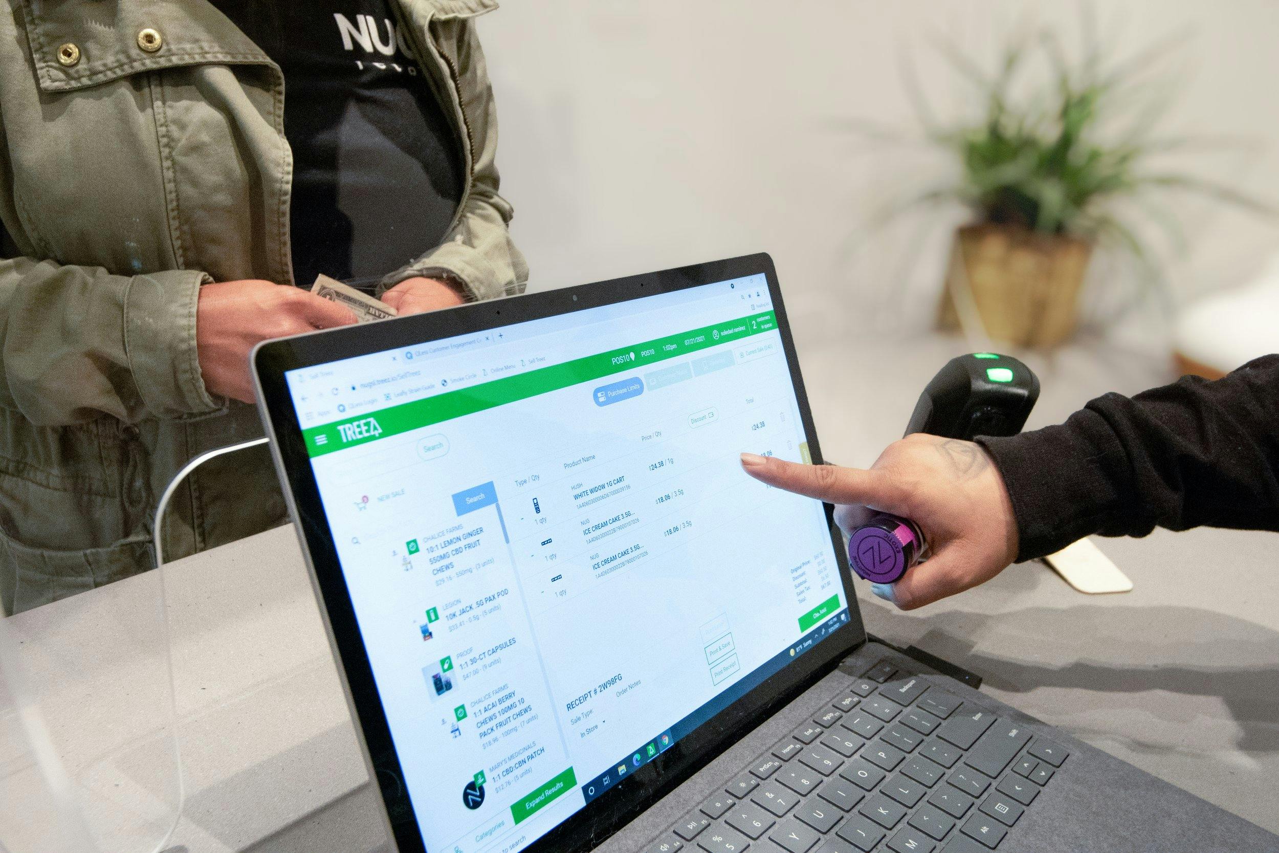 A person waits with cash to purchase cannabis - at the same time, a budtender checks over an order on the cannabis point of sale system and prepares to hand over a preroll and other cannabis products to the customer