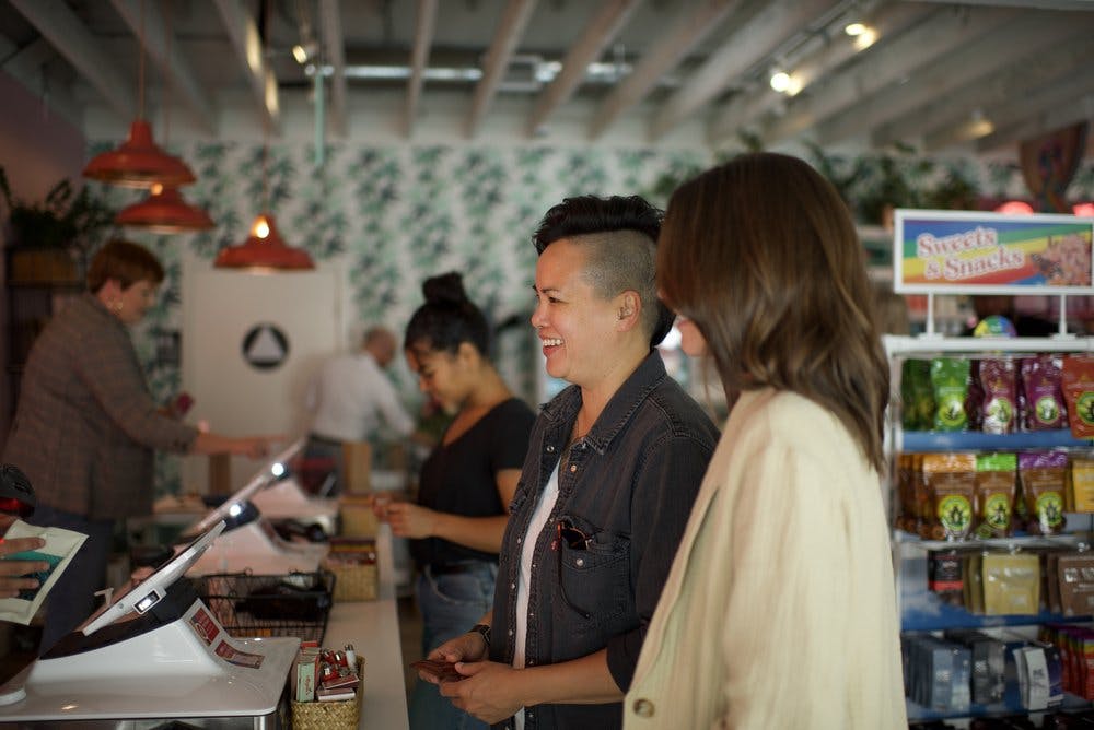 Two customers smile at the check out counter of MOM's Organic Market dispensary. Budtenders are busy helping other customers, and there's a display case of cannabis edibles in the background