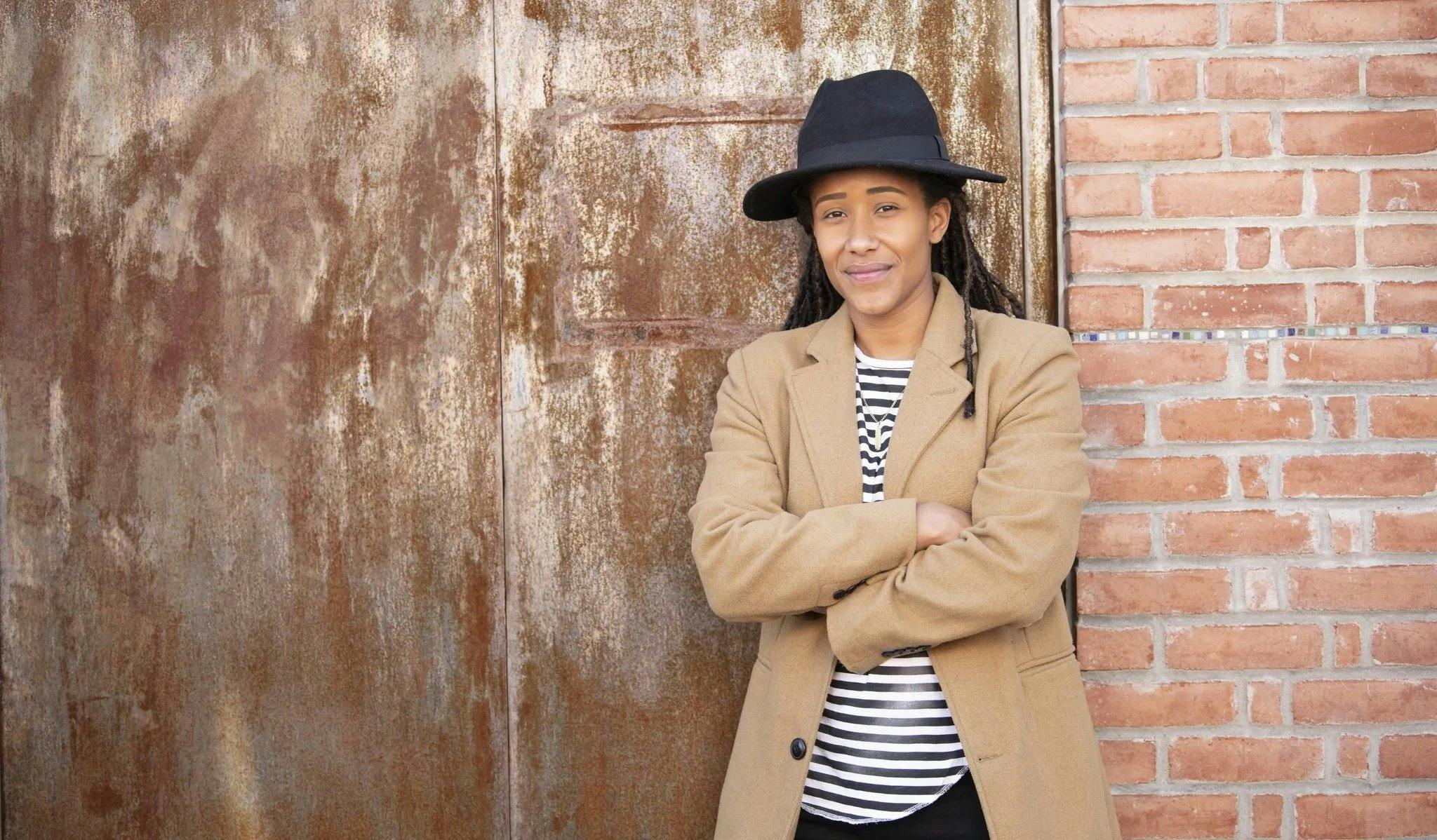 A black woman in a brimmed hat, blazer and striped shirt stands with her arms folded against a brick and metal backdrop. She has a slight smile on her face and looks directly at the camera