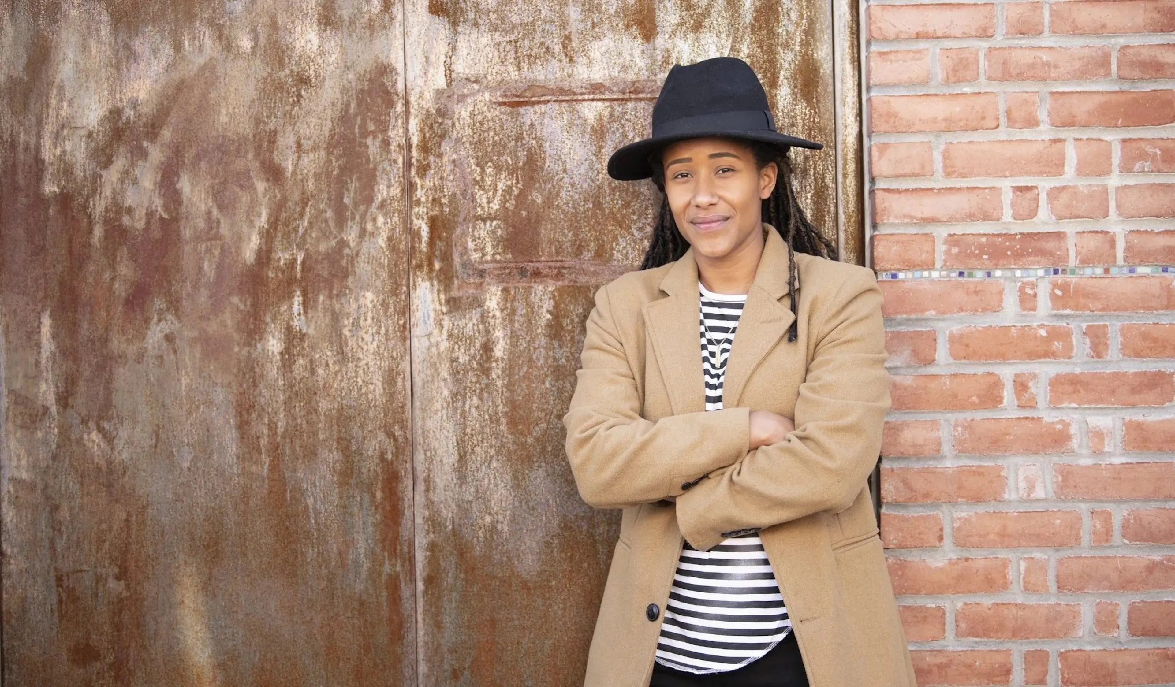 A black woman in a brimmed hat, blazer and striped shirt stands with her arms folded against a brick and metal backdrop. She has a slight smile on her face and looks directly at the camera