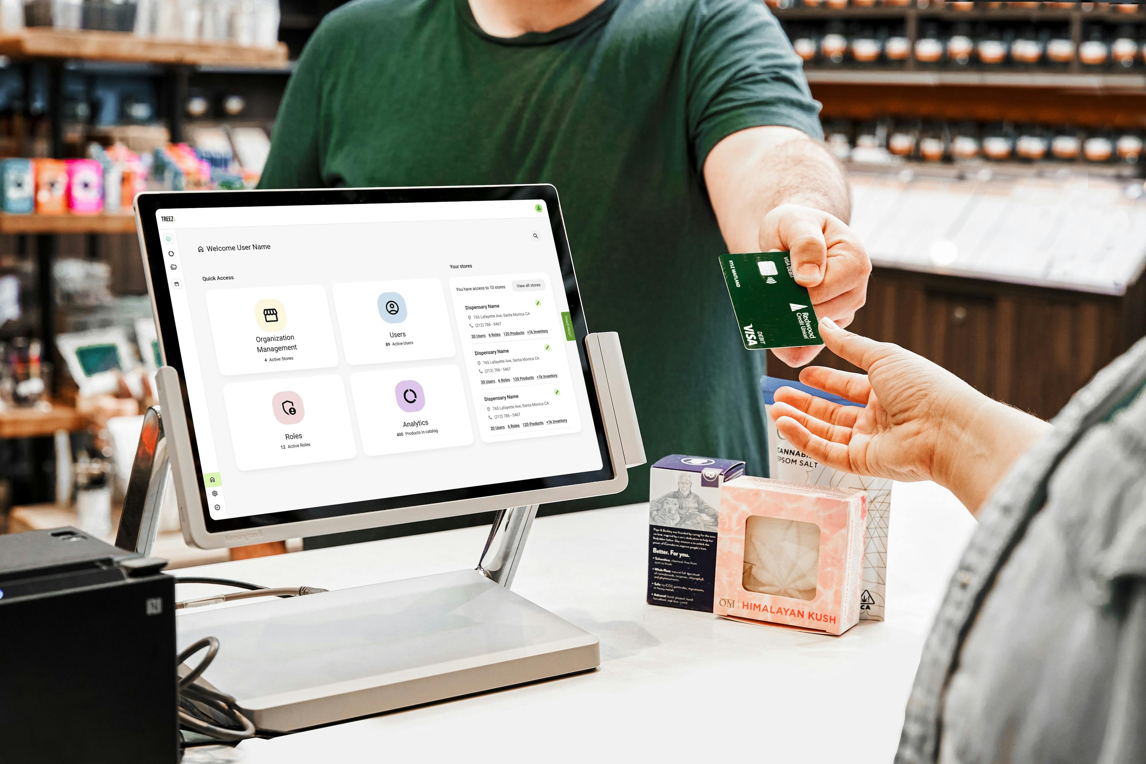 A person passes their debit card to a dispensary employee. A computer displays a POS system with analytics, roles, stores and users. Products including bath bombs and cannabis epsom salt are being purchased.