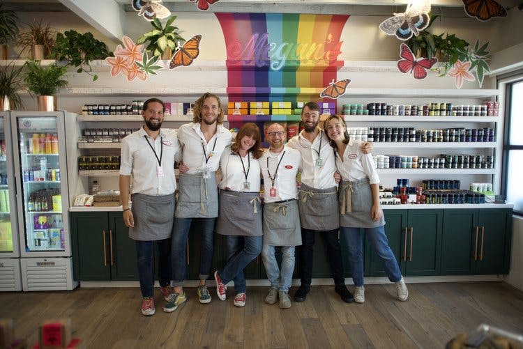 Five staff members of Megan's Organic Market stand in front of a rainbow shelf of cannabis products, to their side is a refrigerator stocked with cannabis beverages