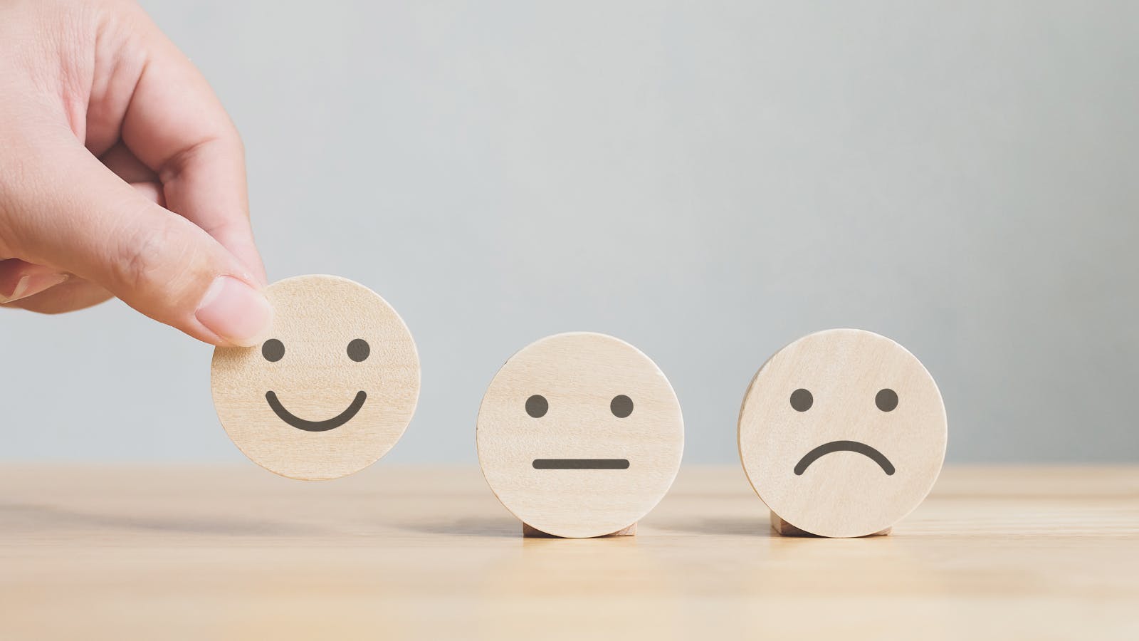 Three wooden smiley face symbols sit on a table - one is a happy face, one an indifferent face with neither a smile nor a frown, and the third face is a sad face