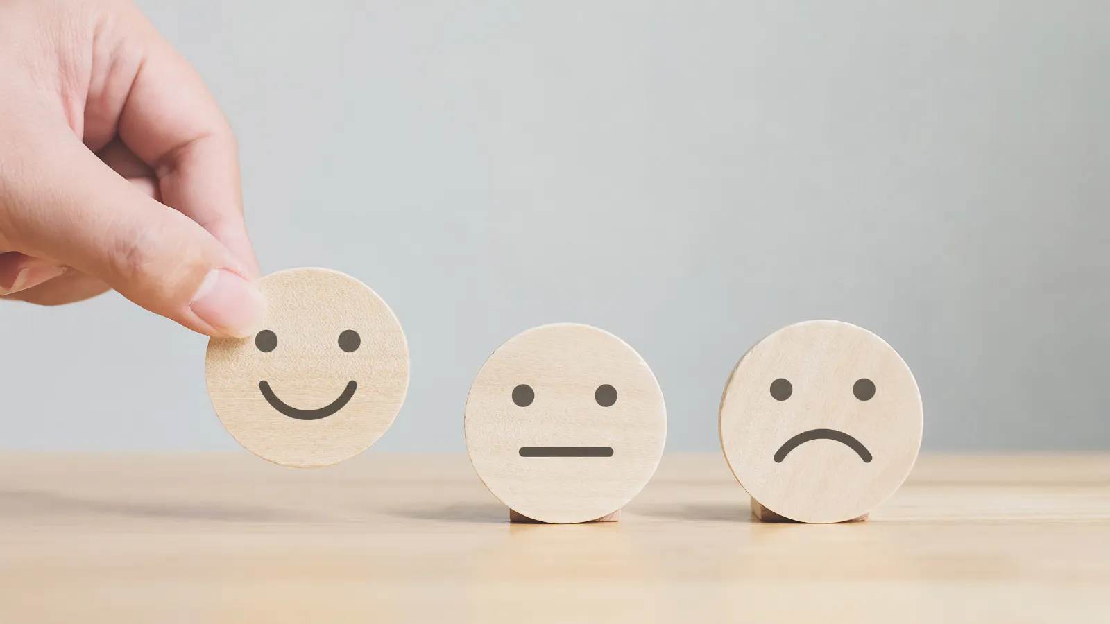 Three wooden smiley face symbols sit on a table - one is a happy face, one an indifferent face with neither a smile nor a frown, and the third face is a sad face