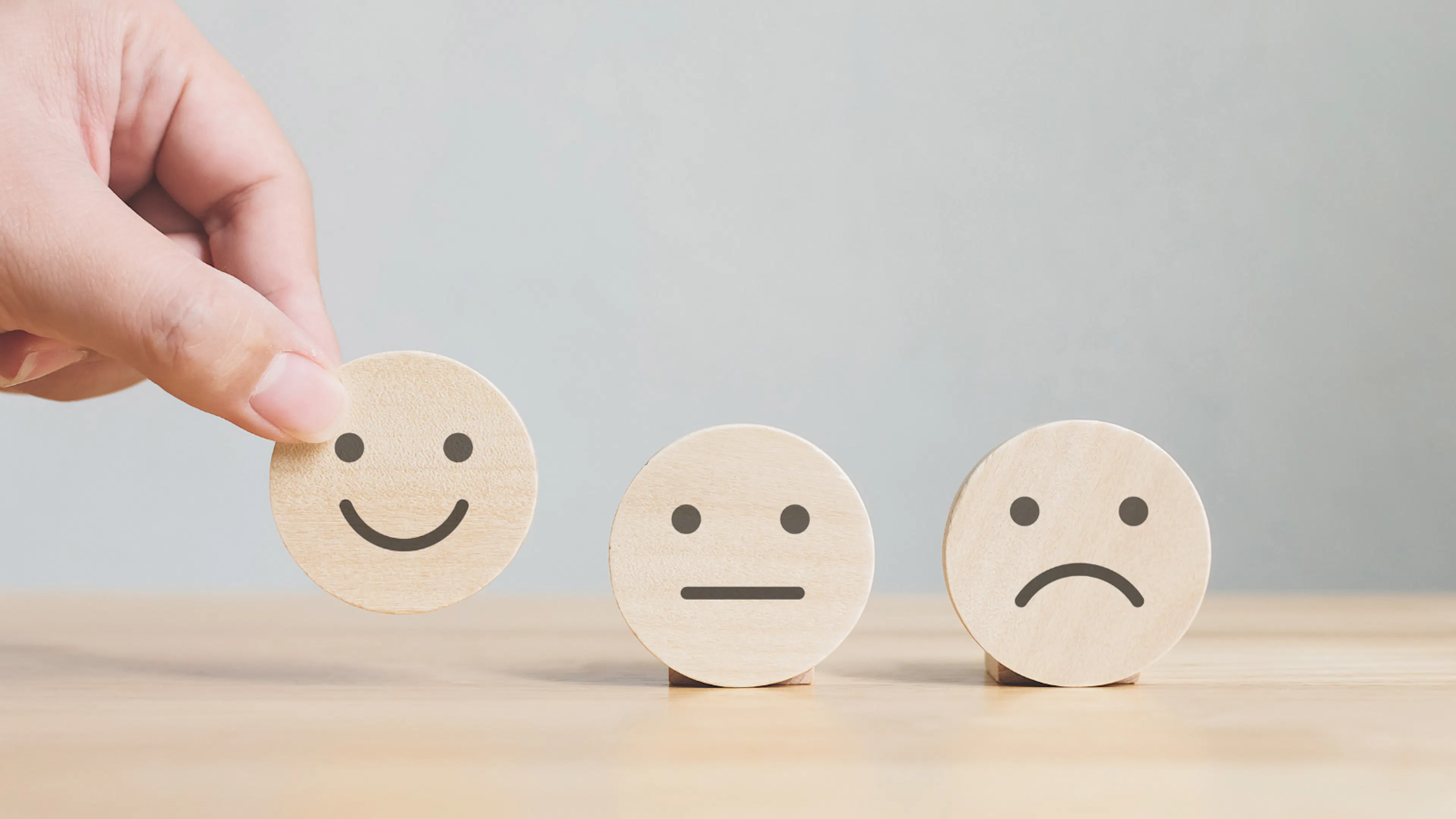 Three wooden smiley face symbols sit on a table - one is a happy face, one an indifferent face with neither a smile nor a frown, and the third face is a sad face