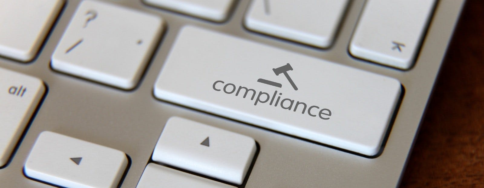A stylized computer keyboard close up with the return button replaced with one that says "compliance" with a symbol of a hammer of justice