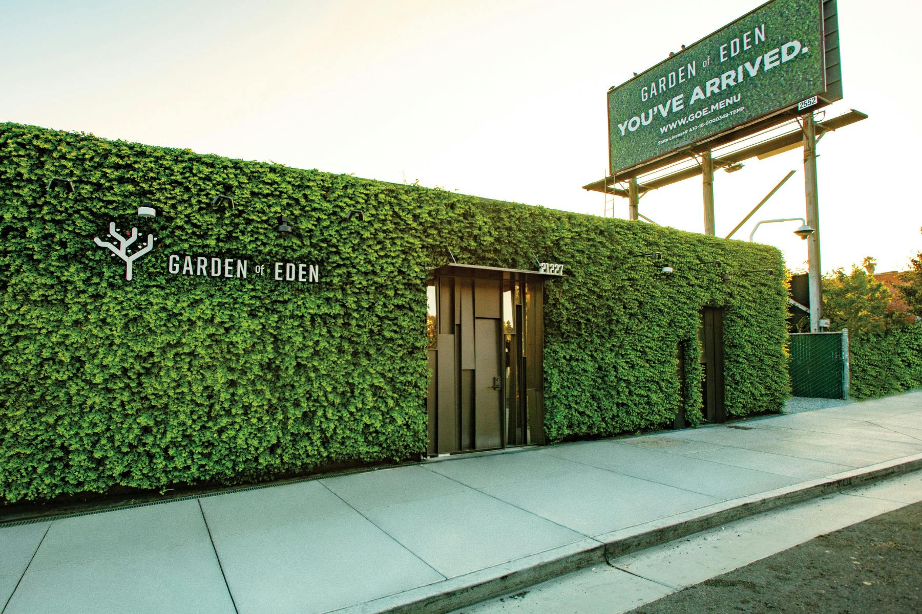 A picture shows the ivy-covered modern cannabis dispensary Garden of Eden bathed in sunset light. Above the store stands a billboard that says Garden of Eden. You've Arrived. www.GOE.menu