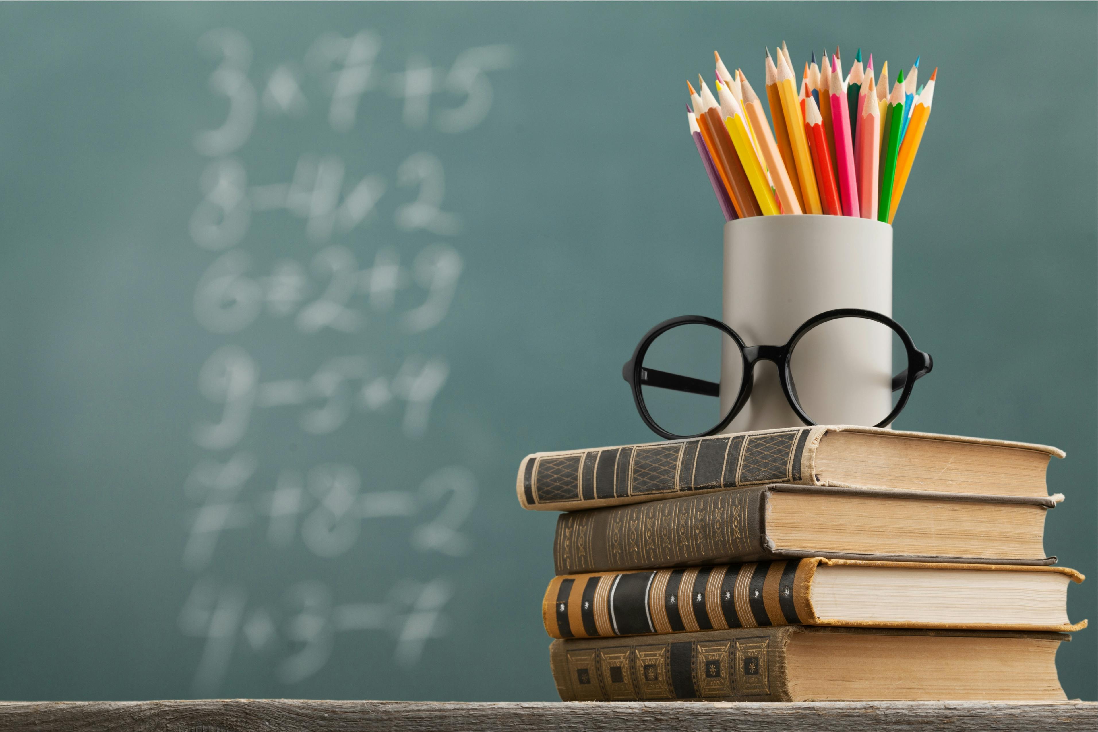 A school scene is shown, with glasses and a cup of pencils stacked on a stack of books, with a chalkboard with math problems in the background