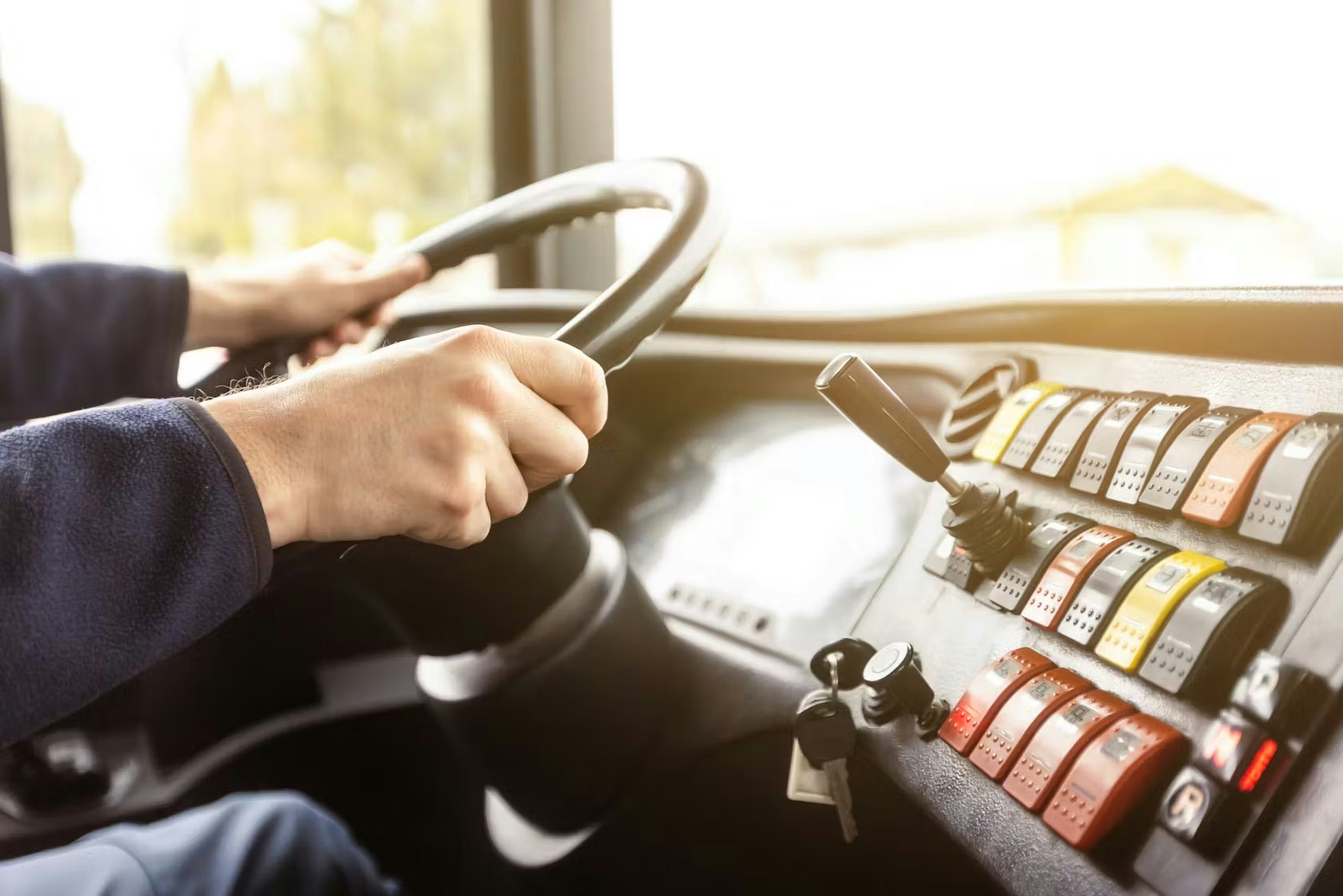 Driver holding a steering wheel inside a vehicle, with dashboard control switches visible.