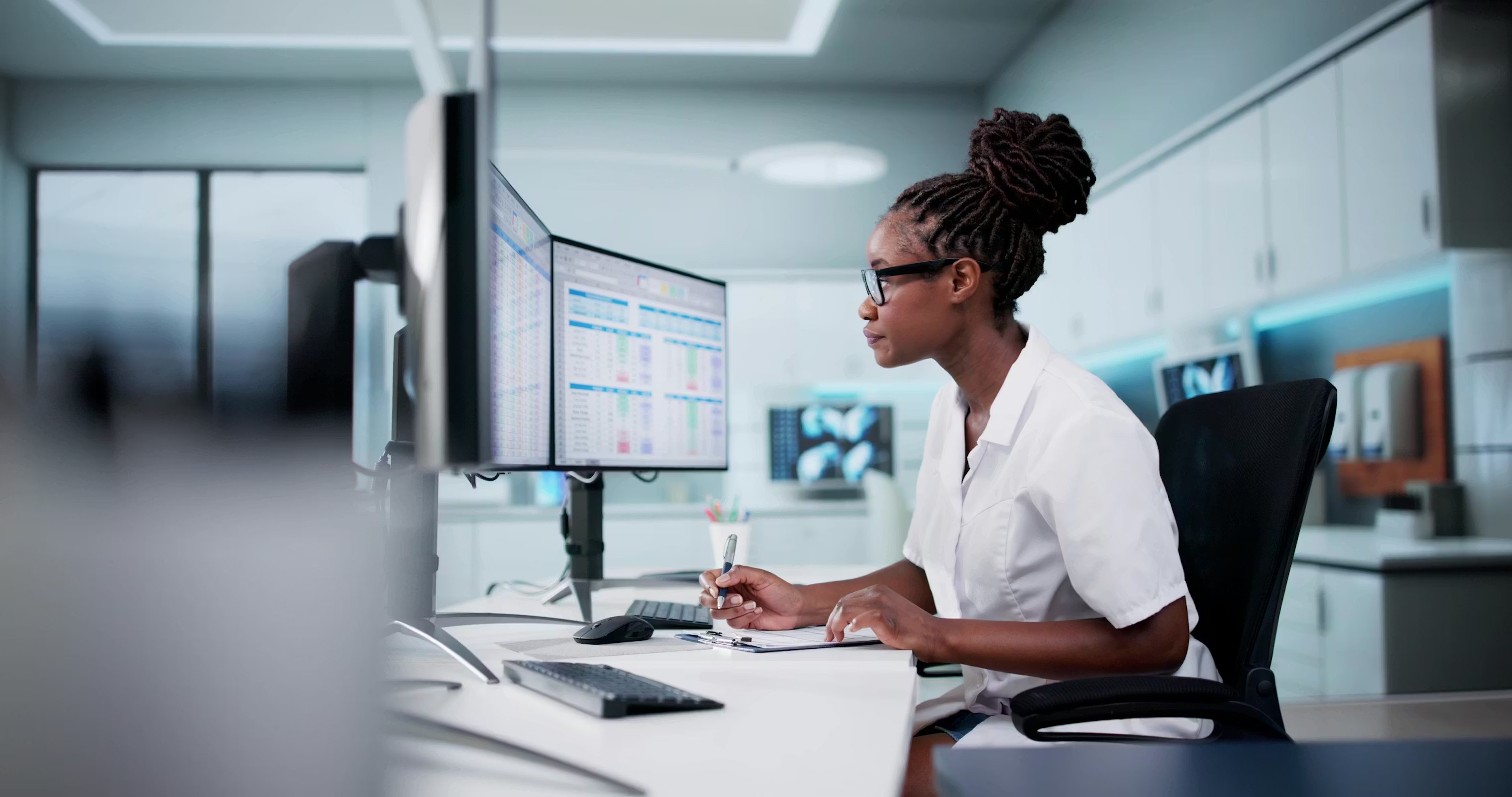 Healthcare professional working at dual computer monitors with spreadsheets in a modern office.