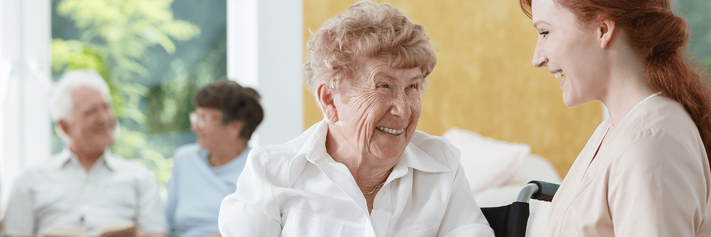 Smiling elderly woman in conversation with a caregiver, with other seniors talking in the background.