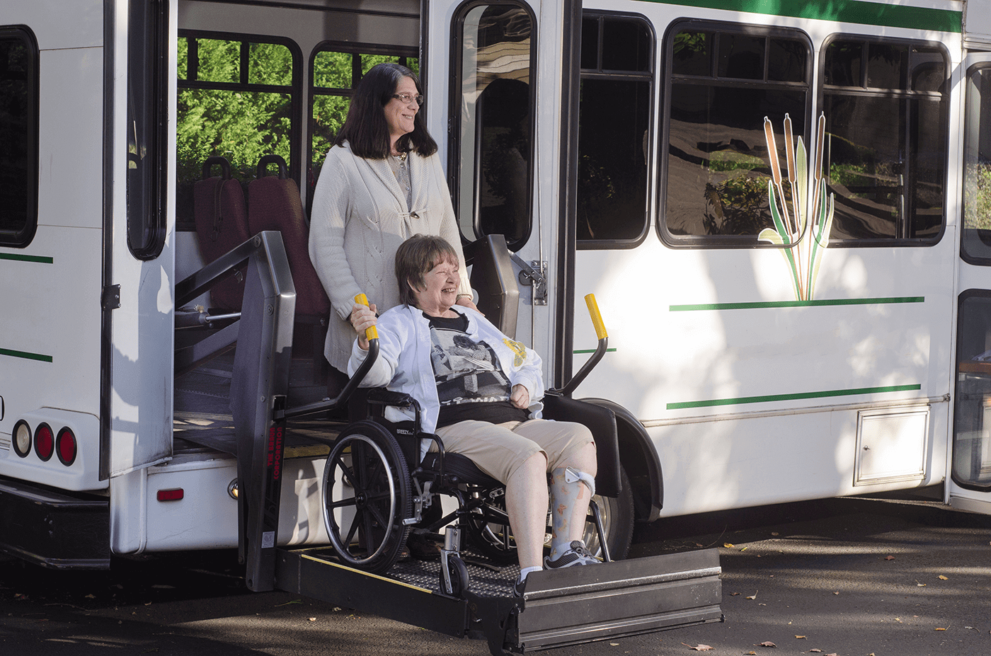 Smiling woman in a wheelchair using a bus lift with assistance from another woman.